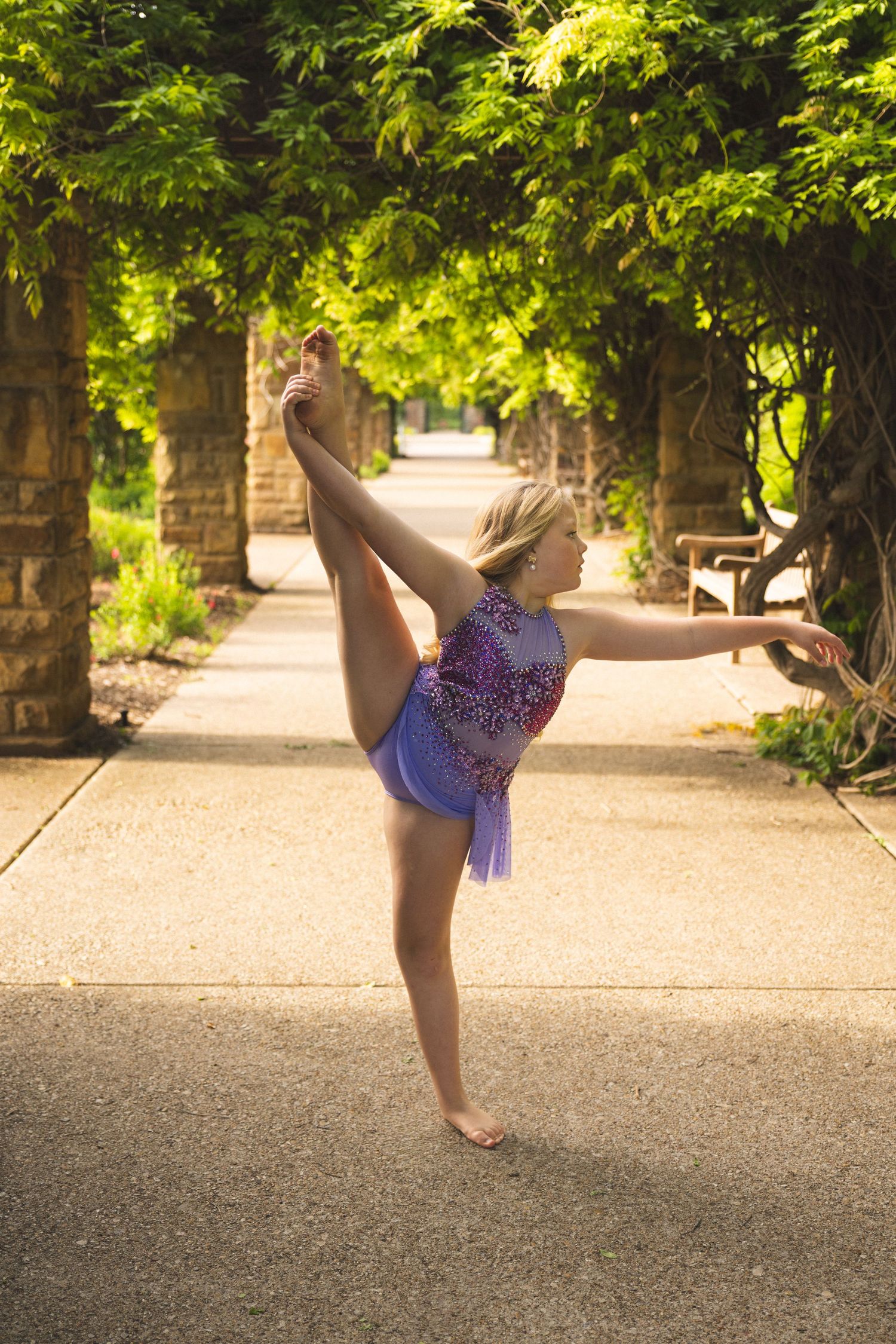 Young gymnast in purple leotard performs a standing split on a tree-lined garden path in warm sunlight.