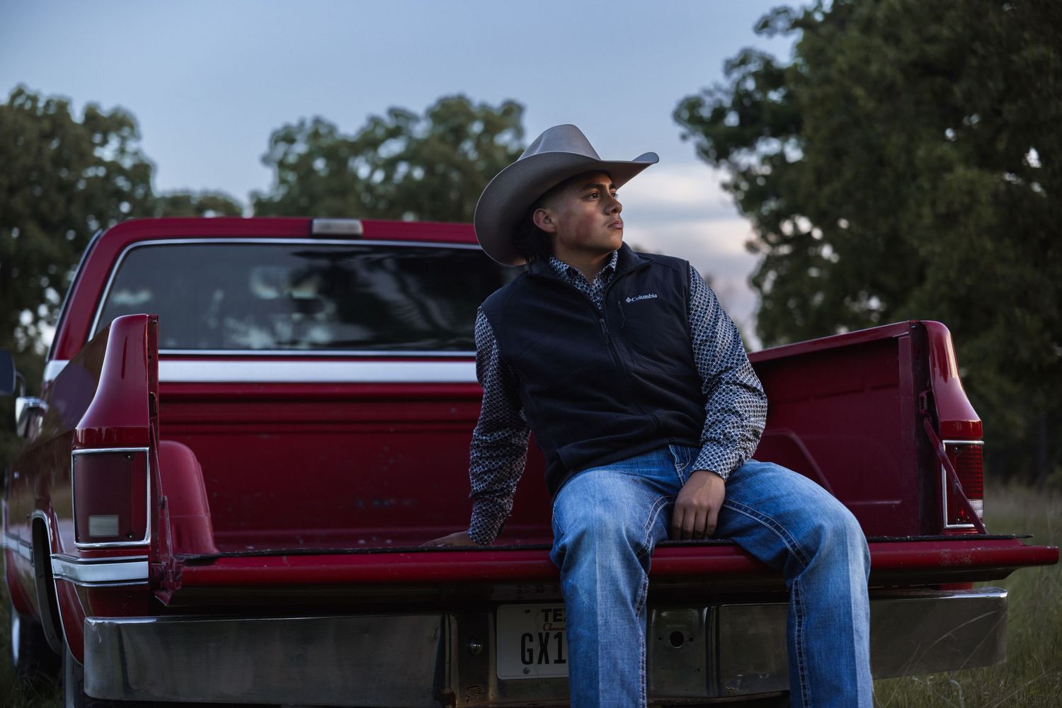 Man in cowboy hat sitting on red pickup truck tailgate in rural setting at dusk.