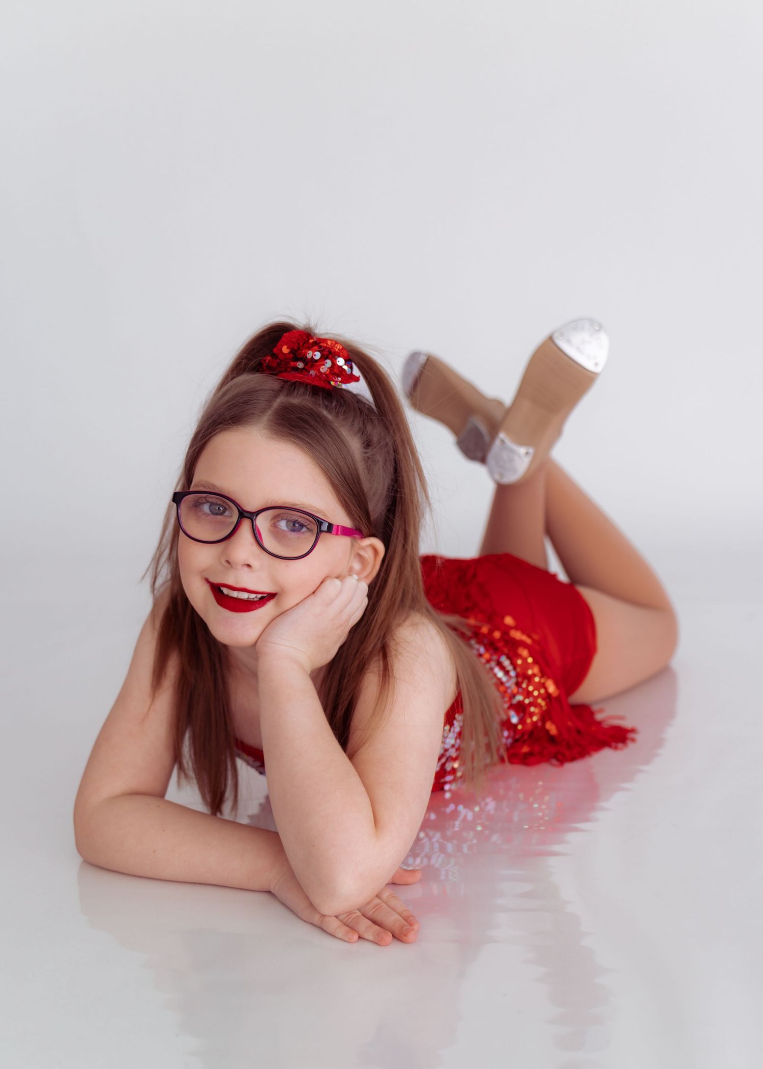 A person in a red dress and glasses lies on the floor in a playful pose against a white background.
