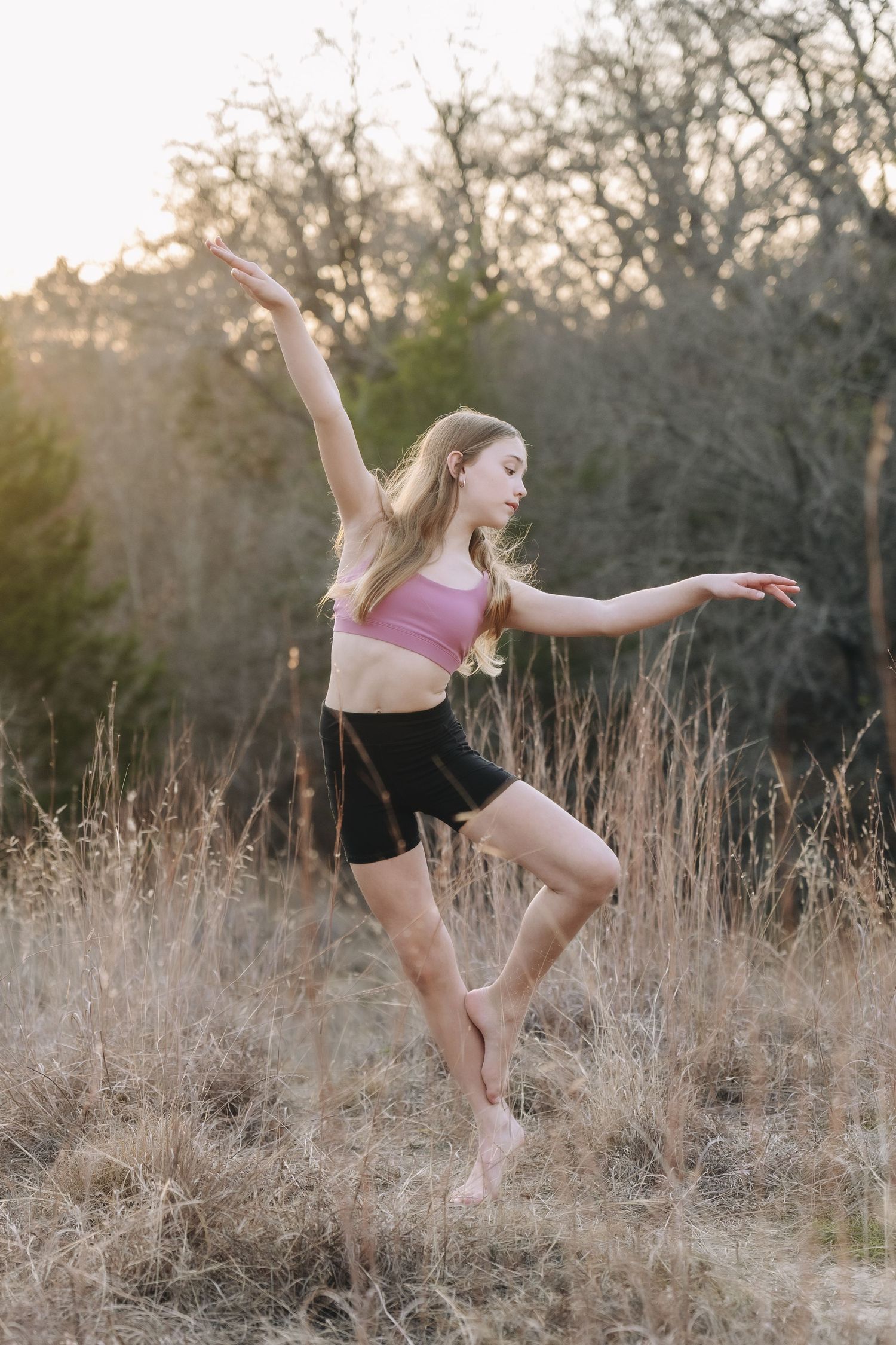 A dancer in pink top and black shorts performs graceful leaps through a golden field at sunset.