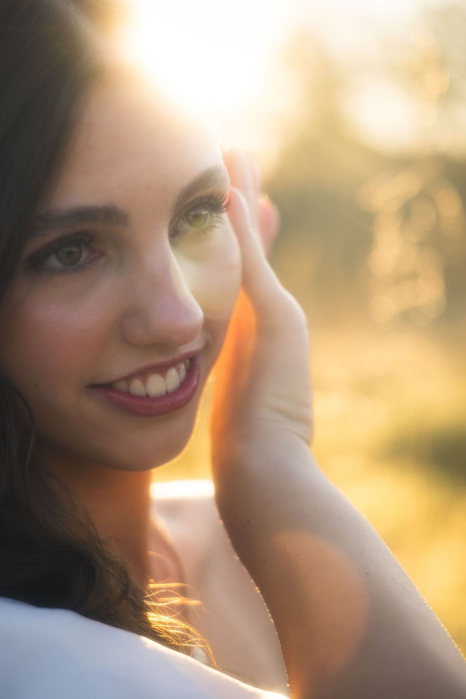 Young woman with dark hair smiling softly in golden sunset light, hand raised near face, bokeh background.