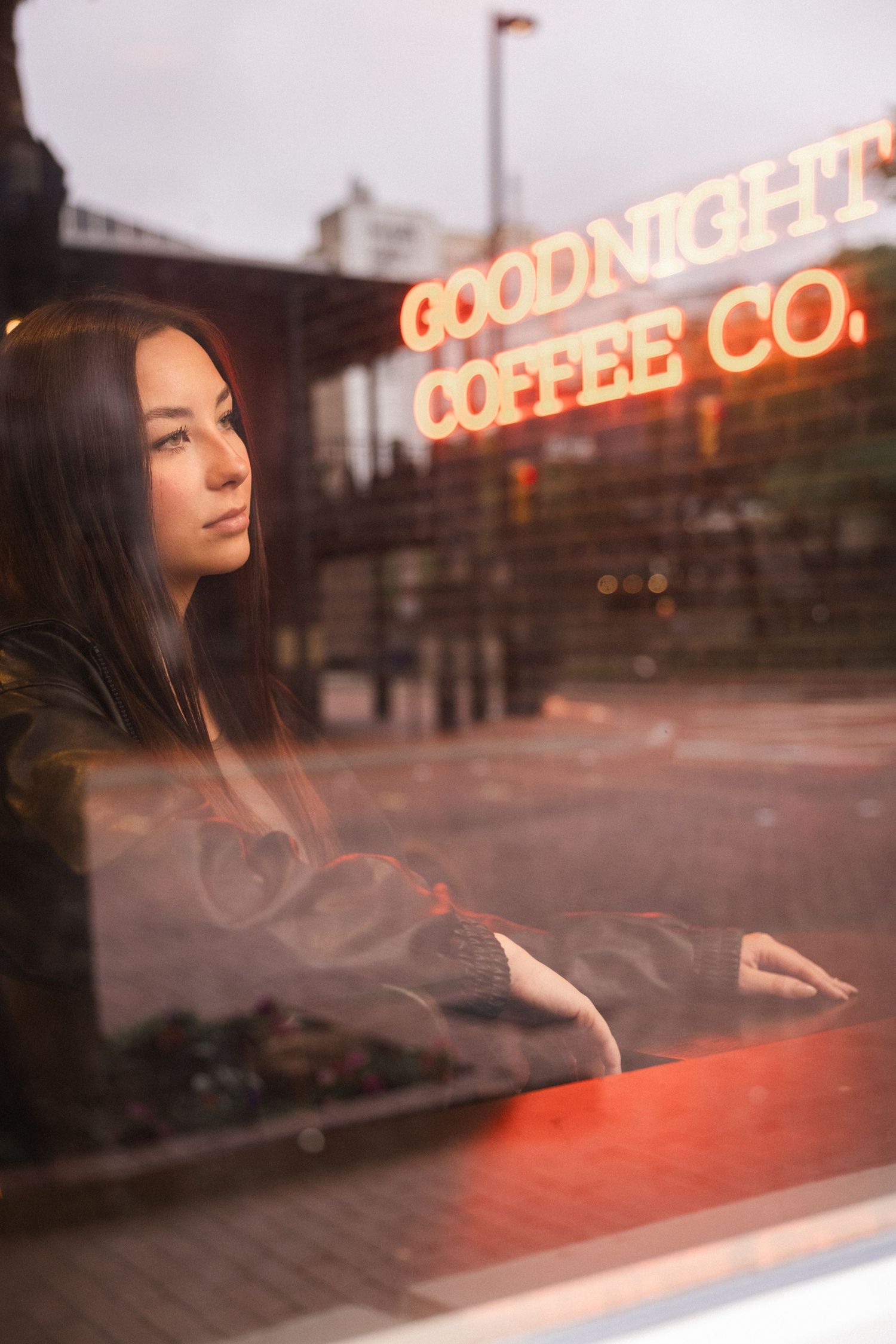 Young woman gazing thoughtfully through a coffee shop window at Goodnight Coffee Co. with neon sign reflection.