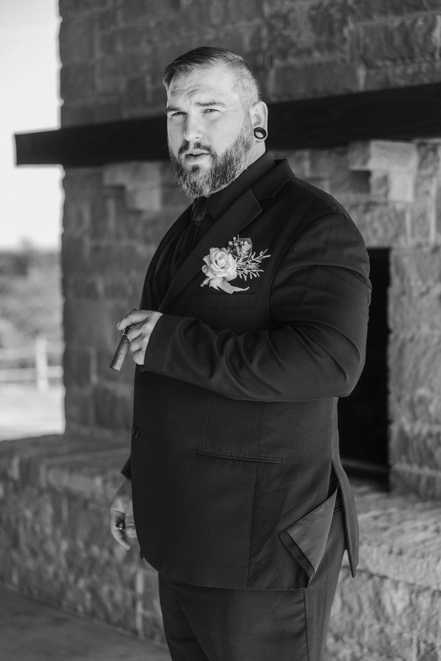 Bearded groom in black suit with floral boutonniere holding cigar in black and white wedding portrait.