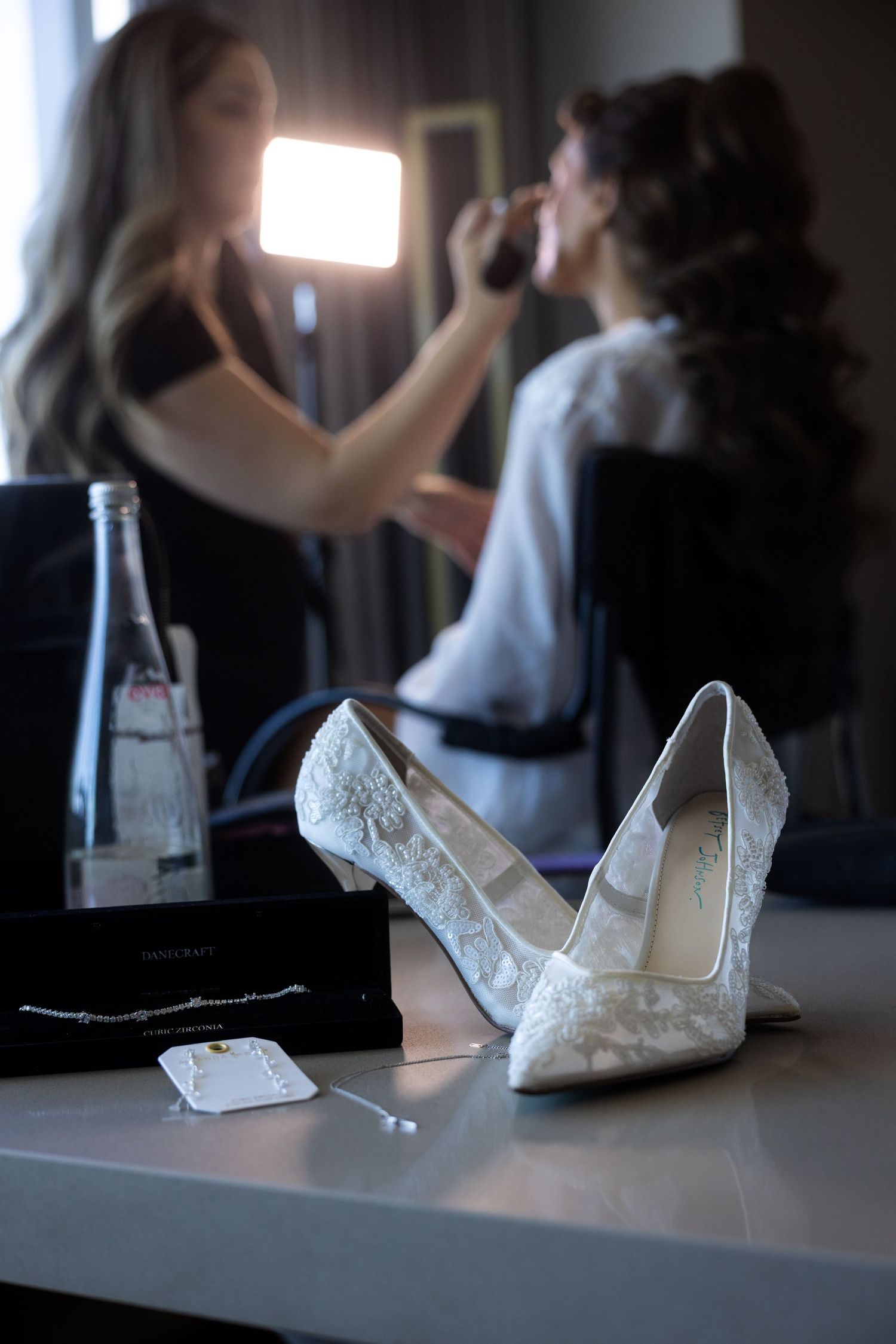 Elegant white lace bridal heels on vanity table with bride getting makeup done in background.