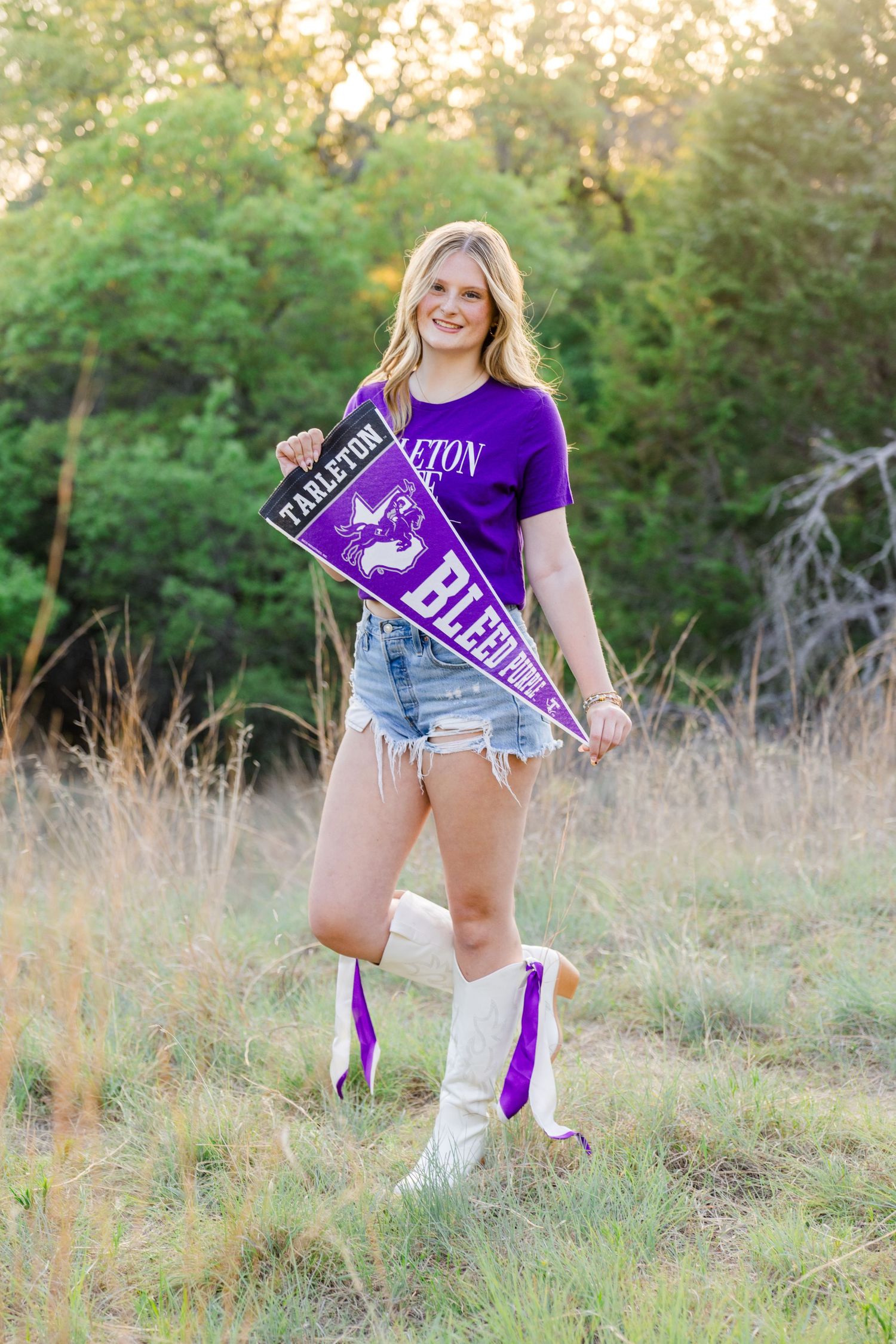 Young woman in purple Tarleton shirt and cowboy boots holding school pennants in a sunny field.
