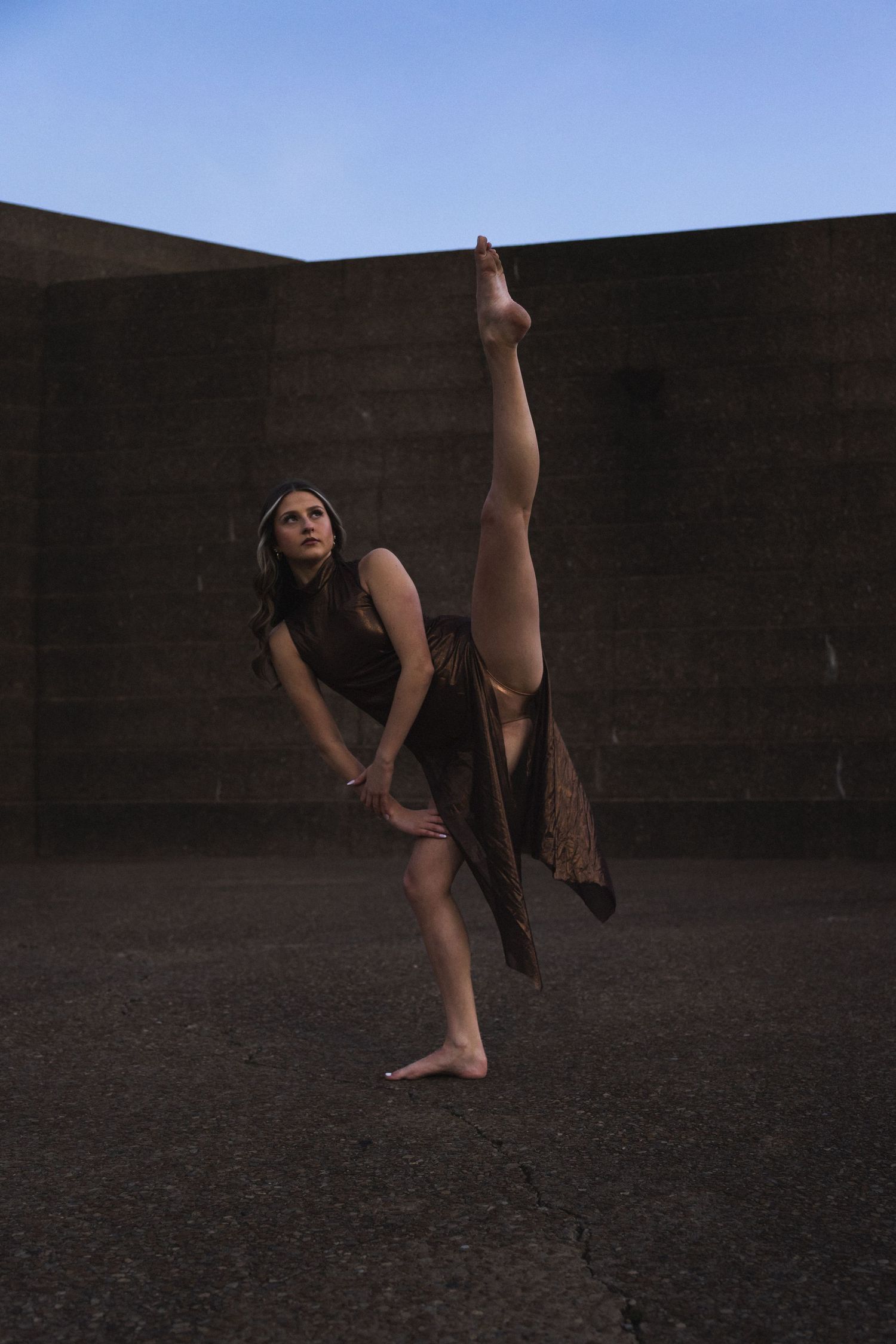A barefoot female dancer performs a high leg extension outdoors against a dark stone wall under a blue sky.