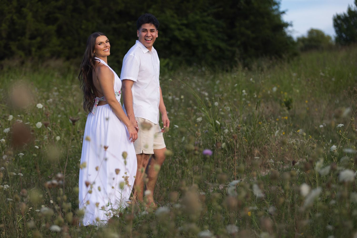 Couple in white outfits holding hands in a wildflower meadow surrounded by lush green trees.