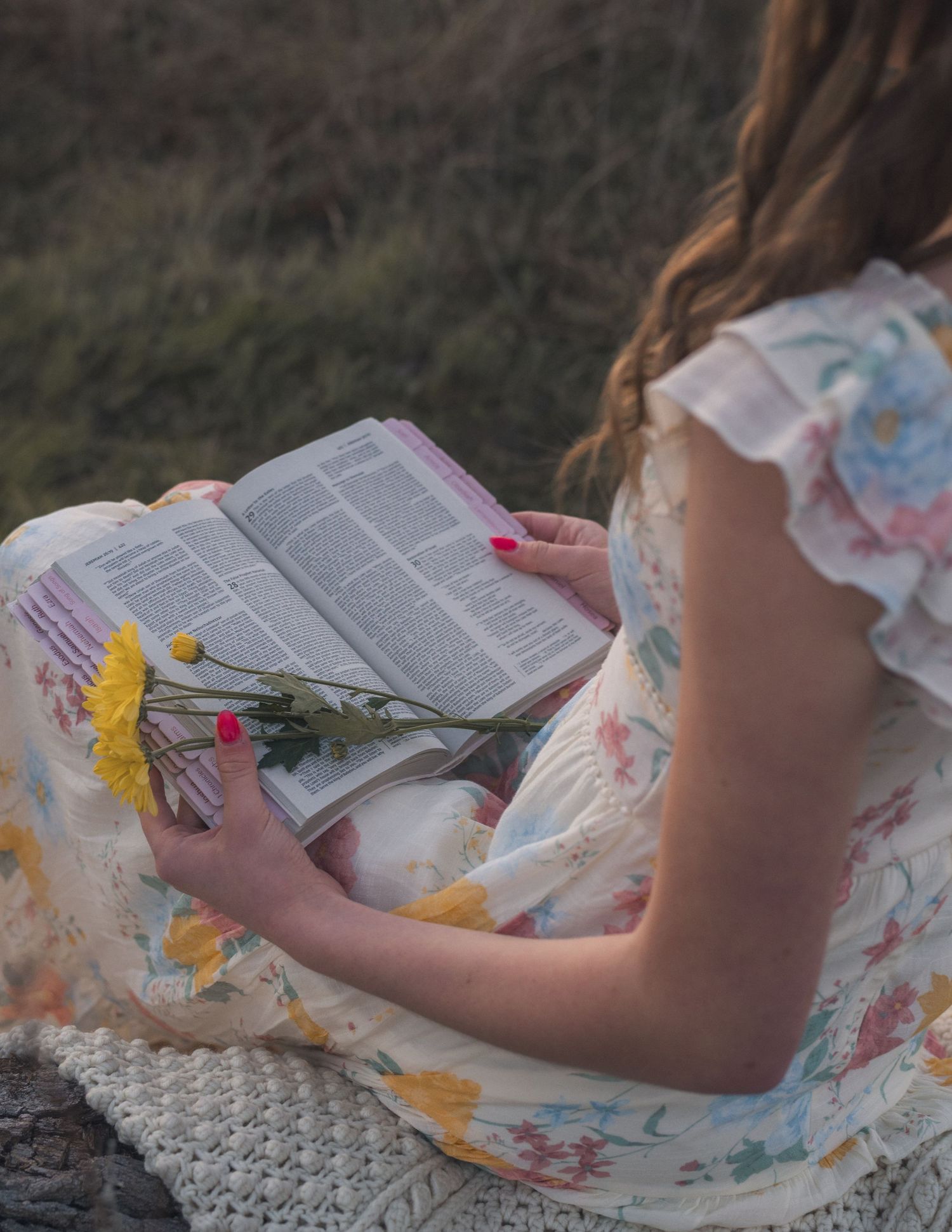 A person in a floral dress sits on a white blanket reading a book outdoors in soft natural lighting.