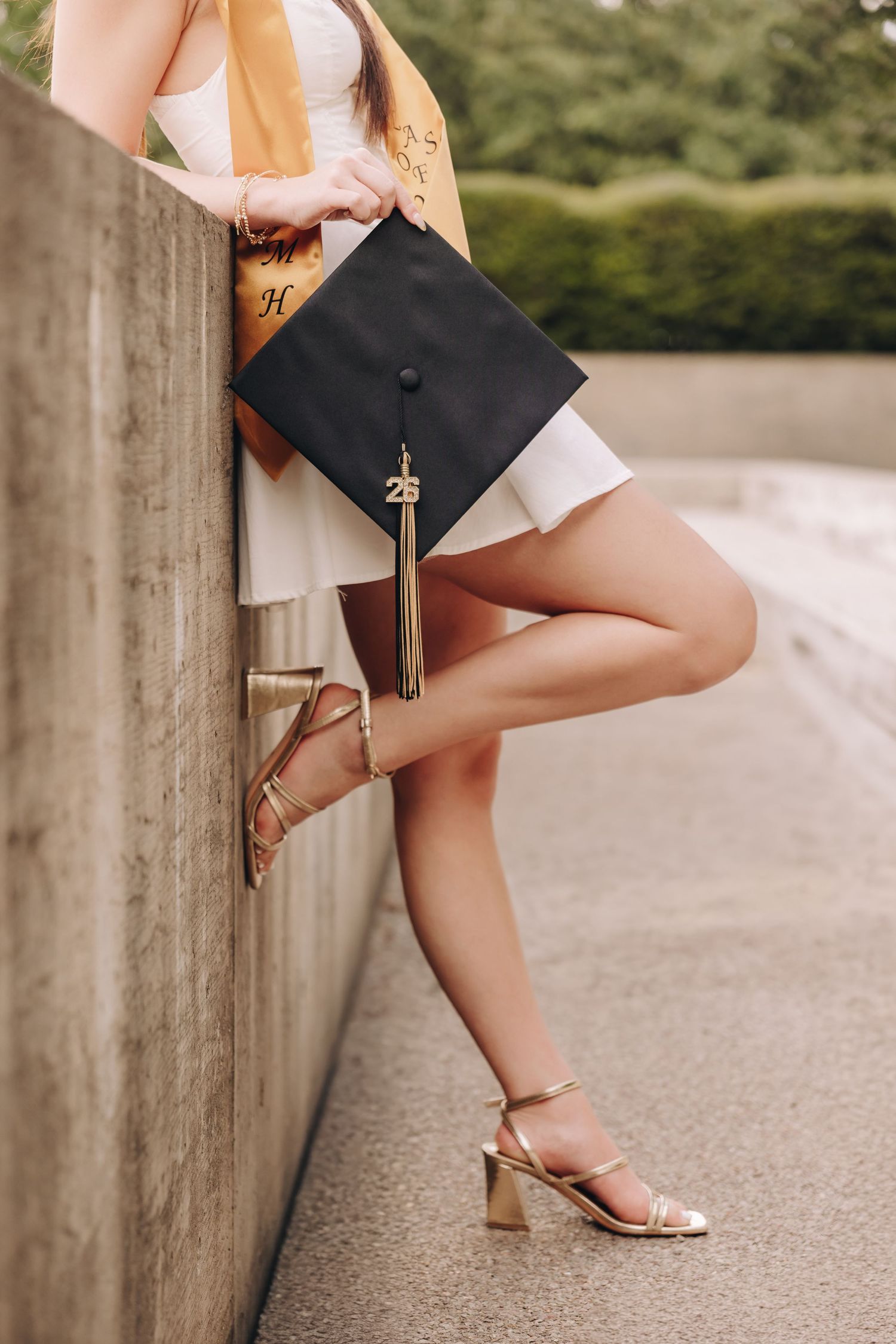A graduate poses in a white dress and gold heels, holding a black graduation cap with tassel outdoors.