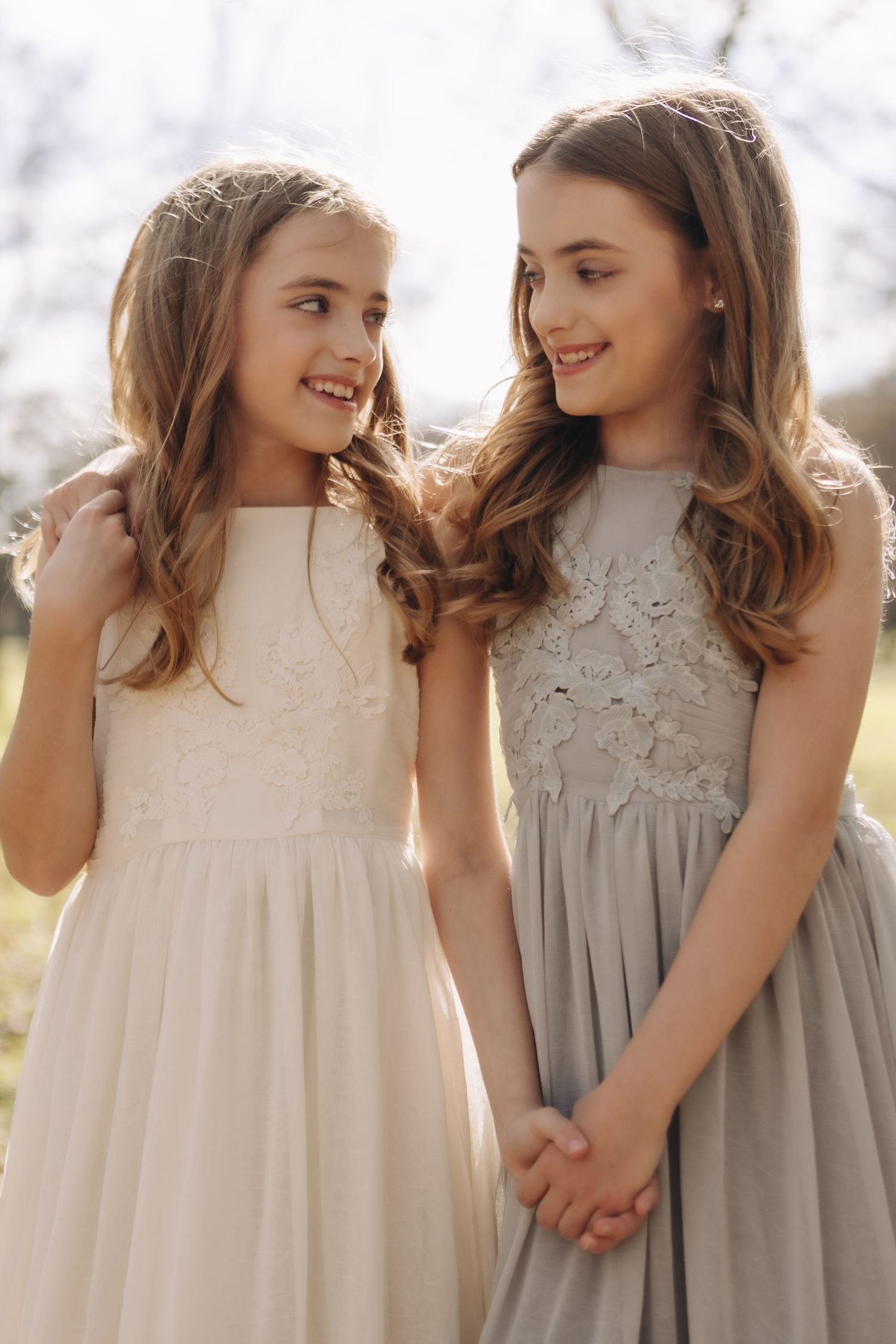 Two young girls in elegant lace dresses hold hands and share a joyful moment outdoors in natural lighting.