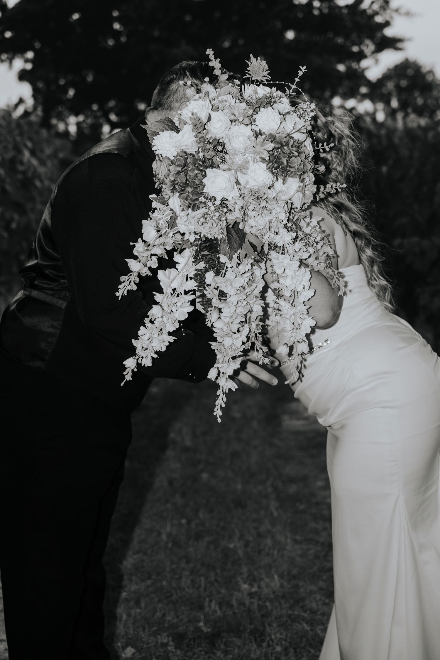 A bride and groom share a kiss hidden behind a lush cascading wedding bouquet of white flowers.