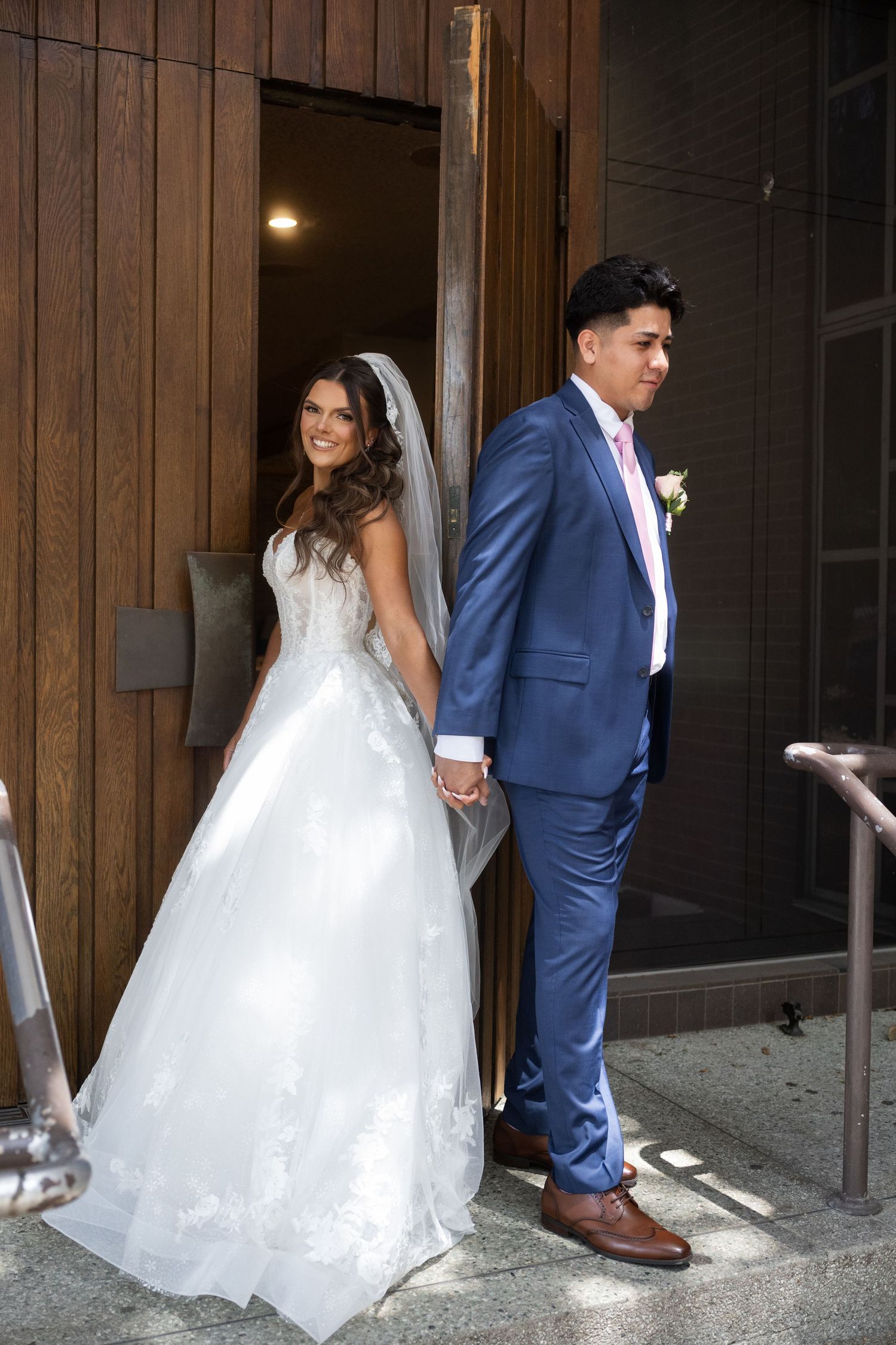 Bride in white lace gown and groom in blue suit holding hands outside wooden doors on their wedding day.