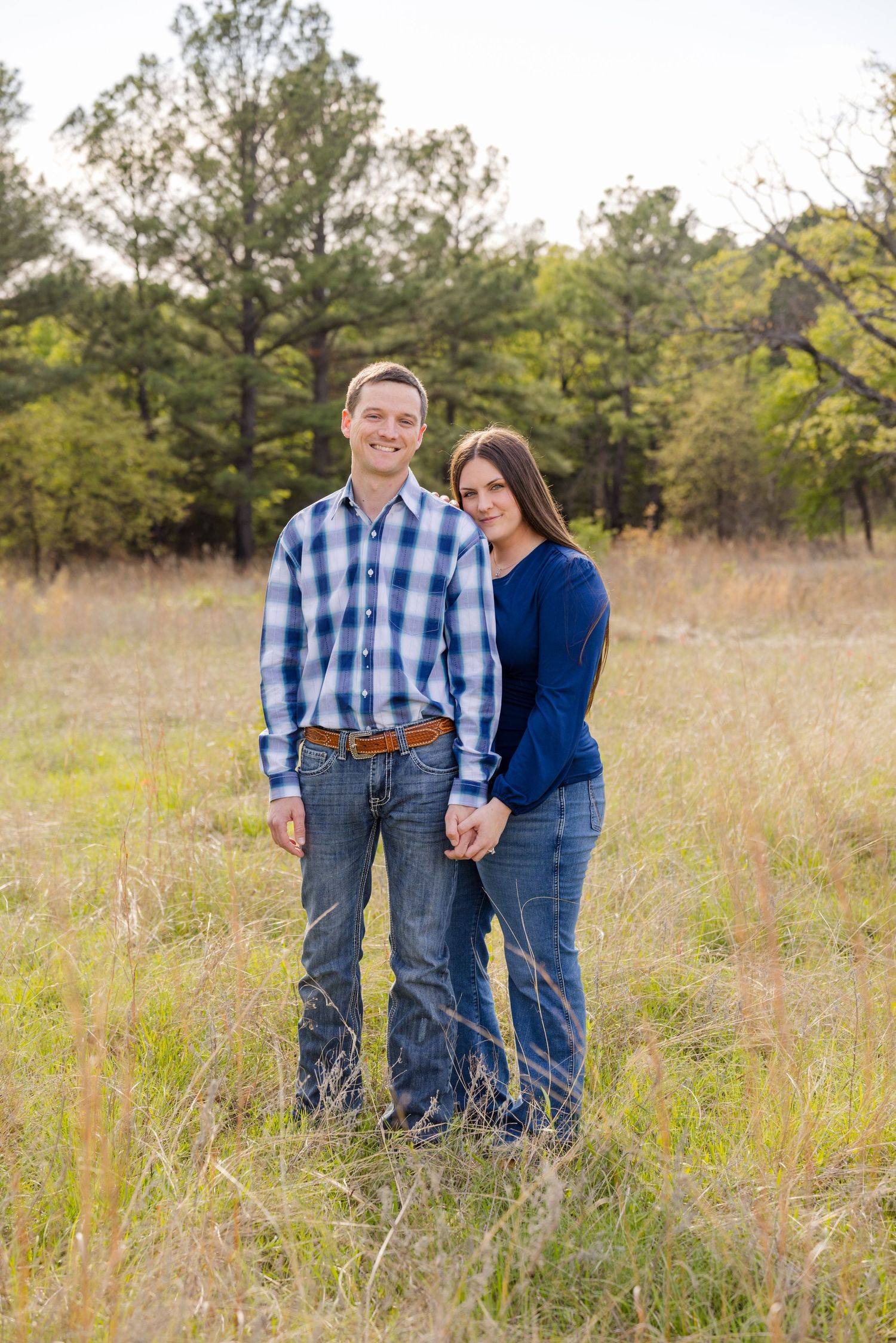A smiling couple holds hands in a sunlit grassy field with pine trees, wearing blue plaid and navy outfits.