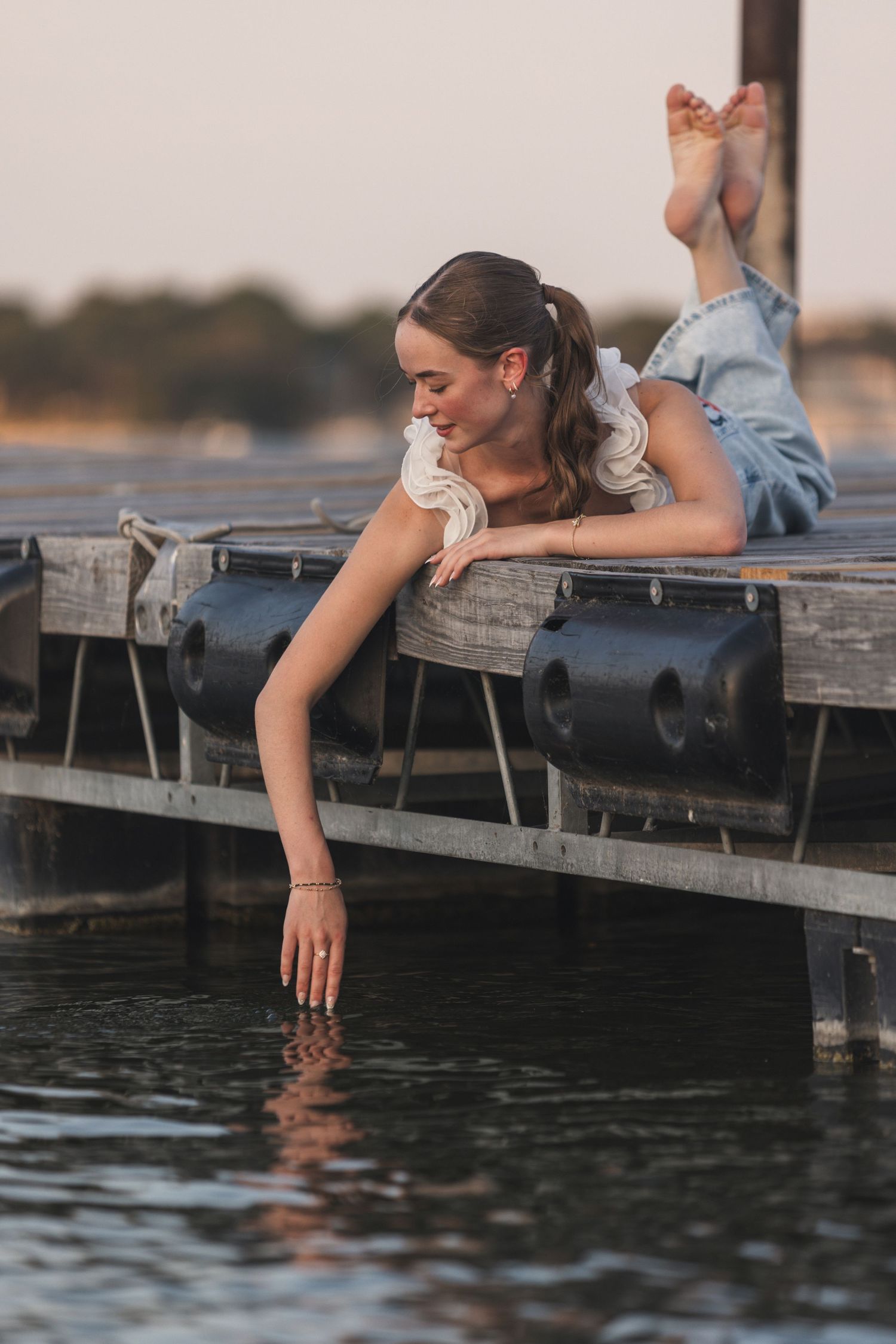 Young woman in denim overalls lying on a wooden dock, touching the calm water below at sunset.