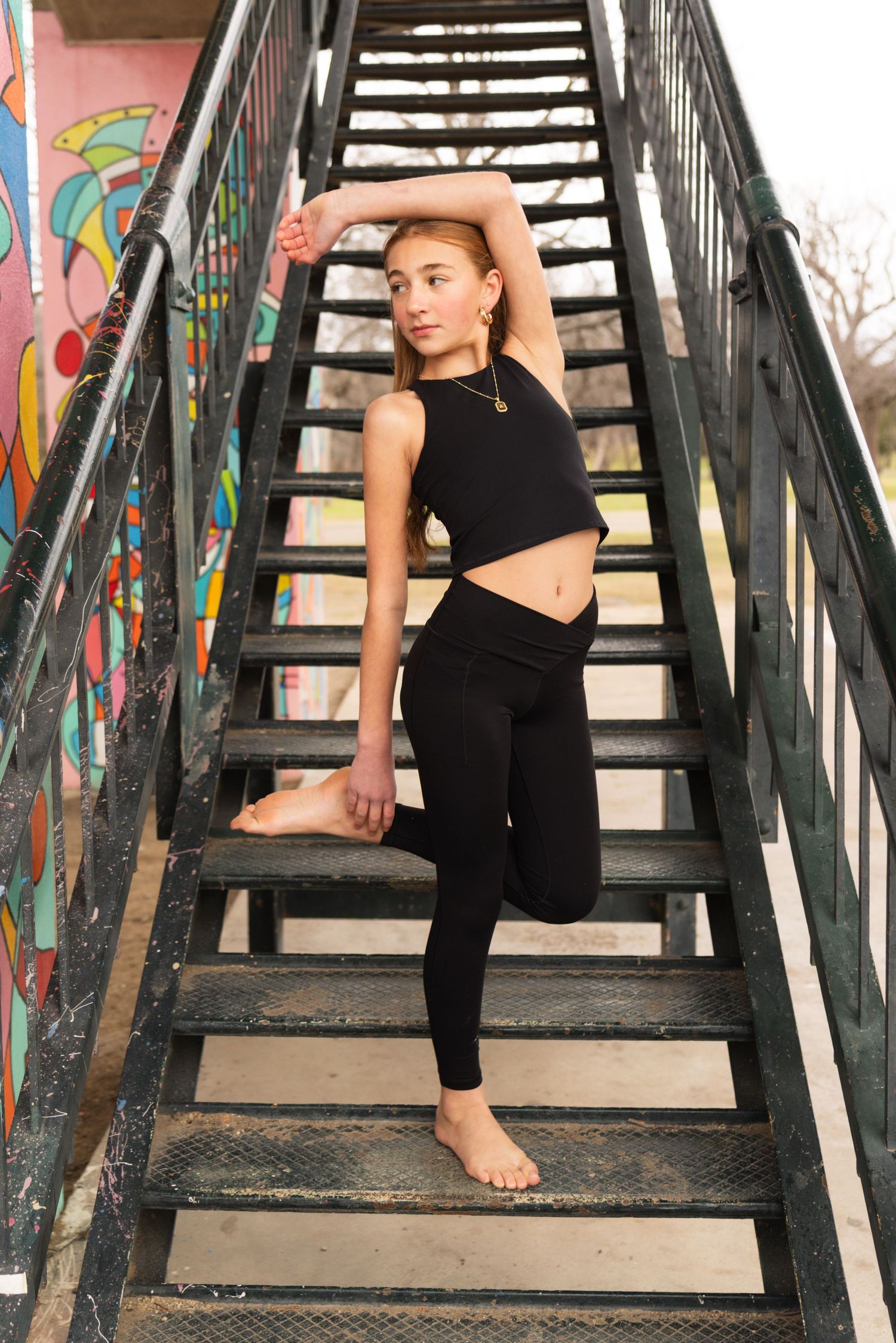 Athletic figure in black workout attire poses on urban staircase with colorful graffiti wall in background.