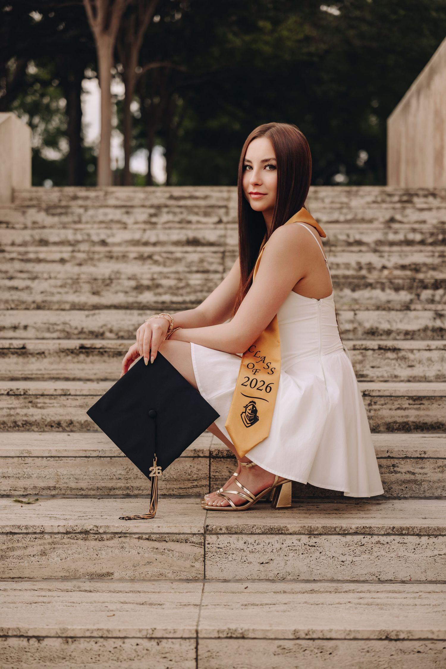 Young woman in white dress holding graduation cap, wearing Class of 2026 sash, sitting on stone steps outdoors.