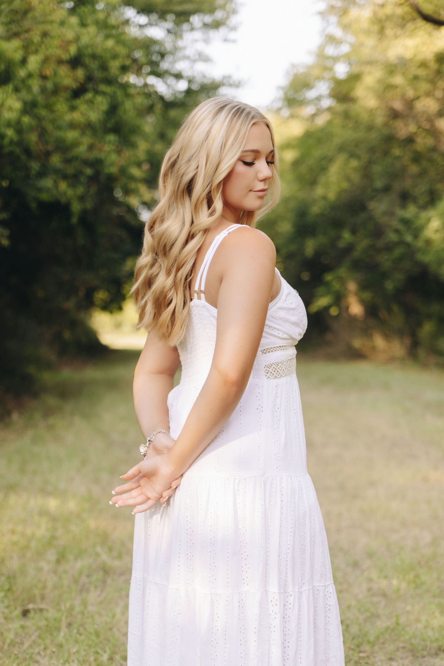 Young woman with blonde wavy hair wearing a white maxi dress standing outdoors in a green park setting.