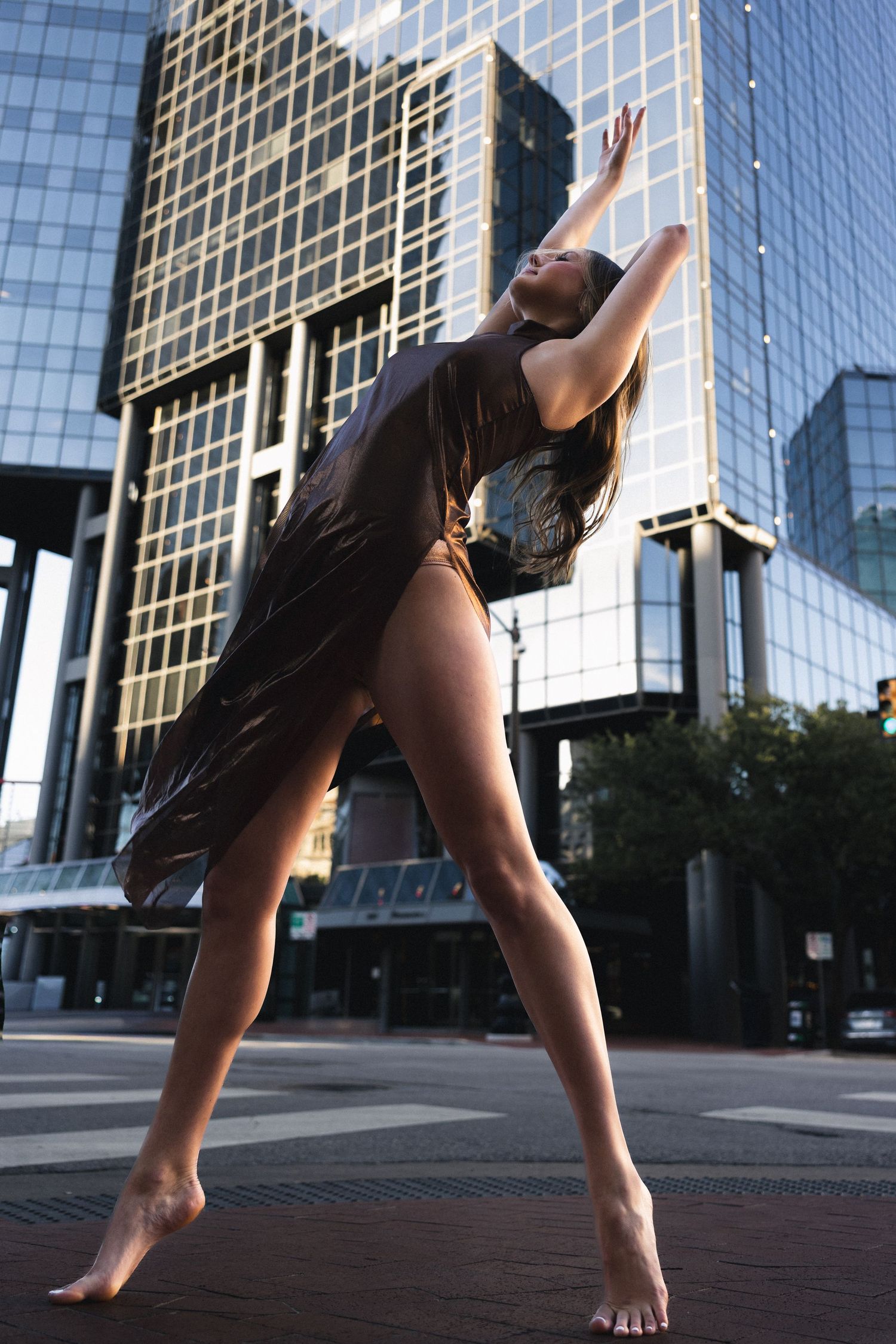 Barefoot dancer poses gracefully on urban street with modern glass skyscrapers in background.