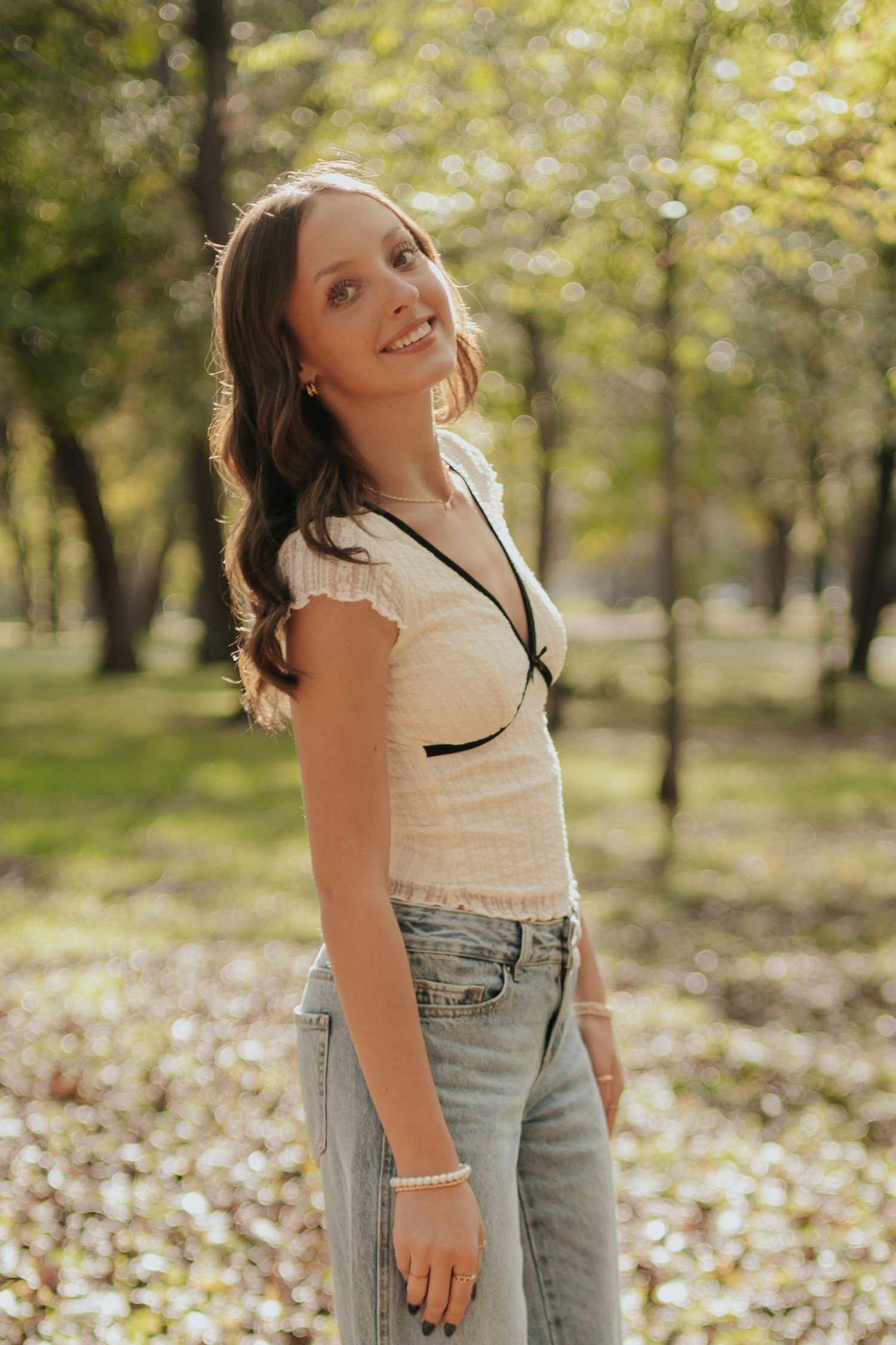Person in light top and jeans poses playfully in autumn park with fallen leaves and trees in background.