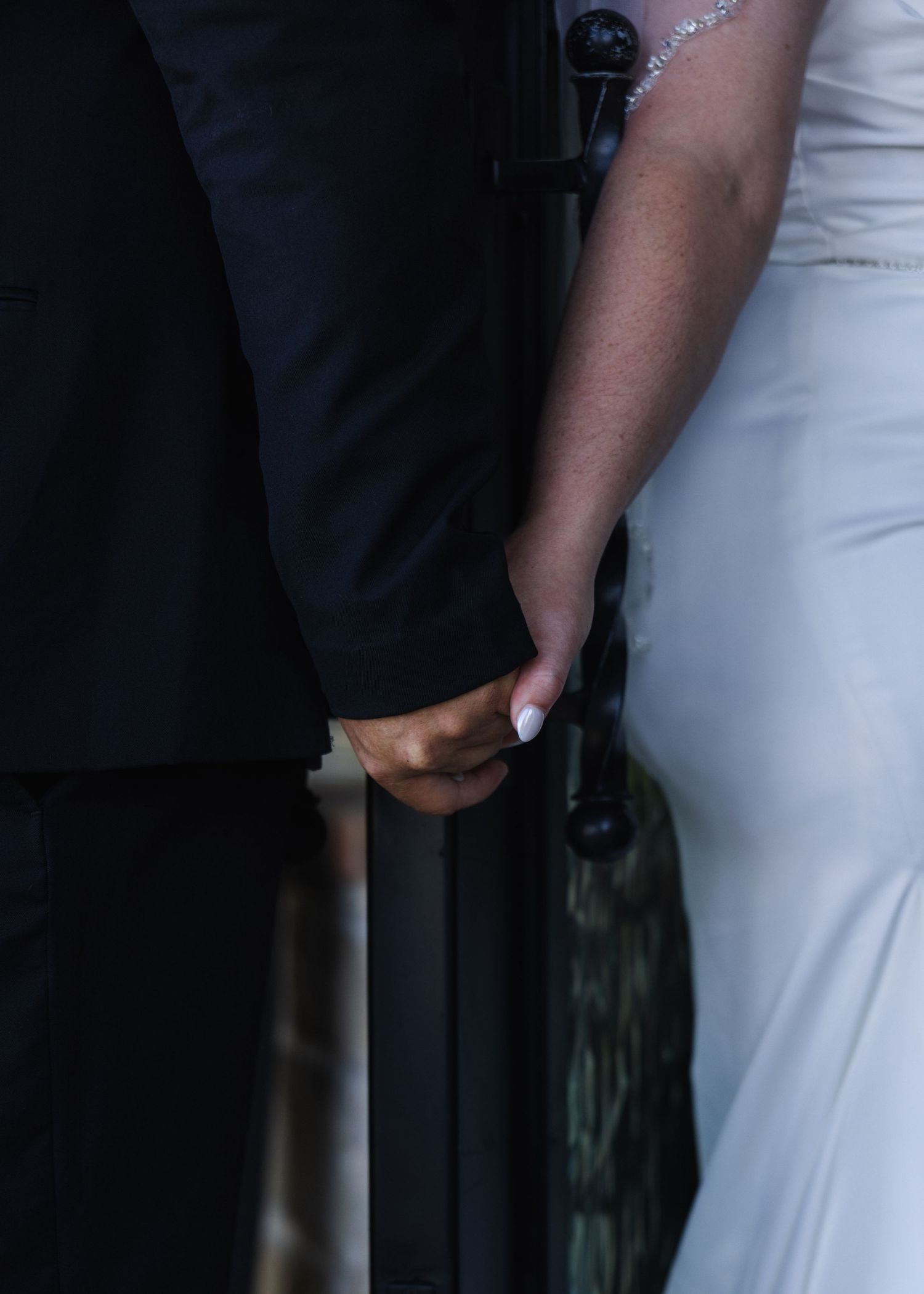 Couple holding hands at wedding, groom in black suit and bride in white gown, close-up detail shot.