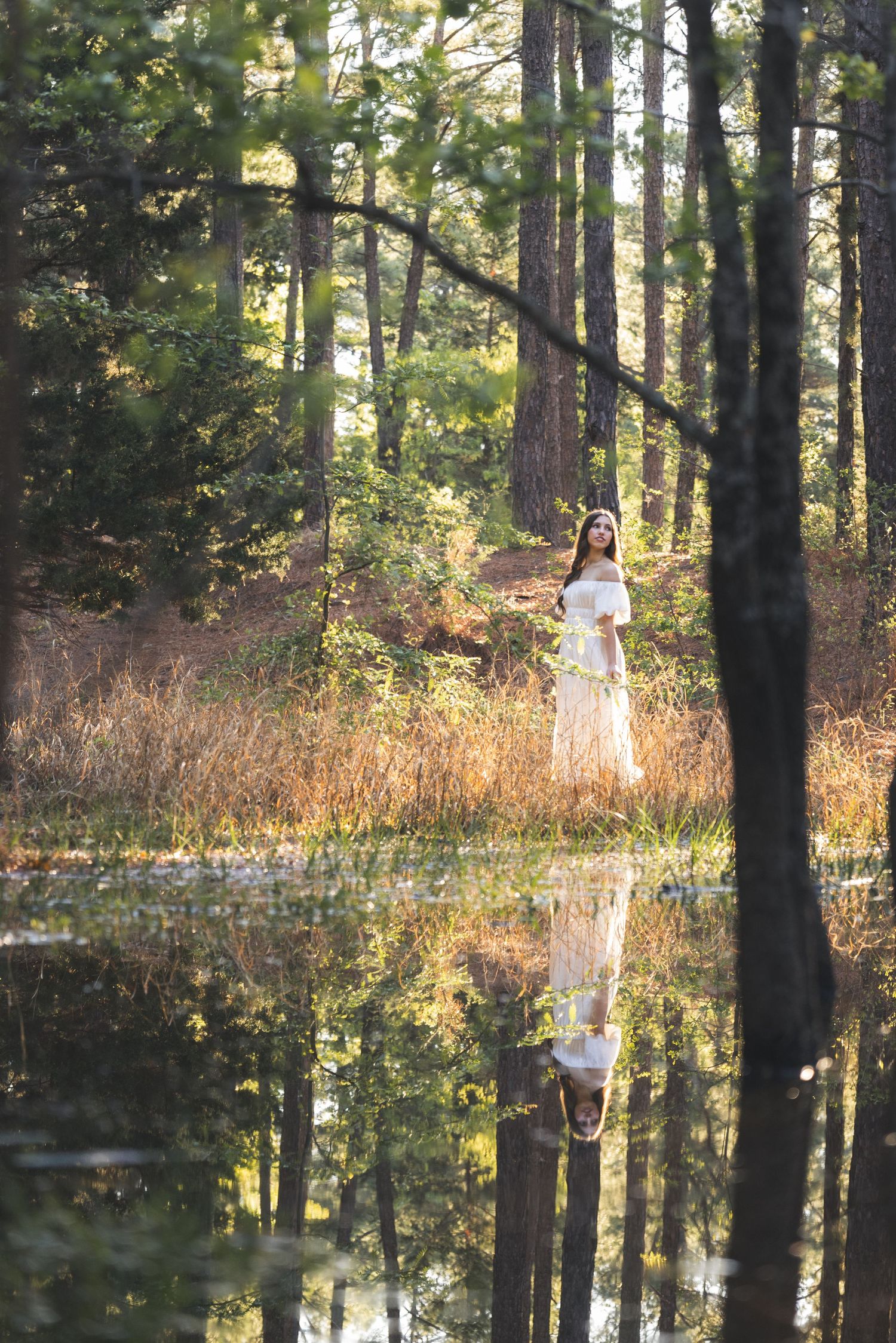 Woman in white dress standing by a reflective forest pond surrounded by tall pine trees in golden light.