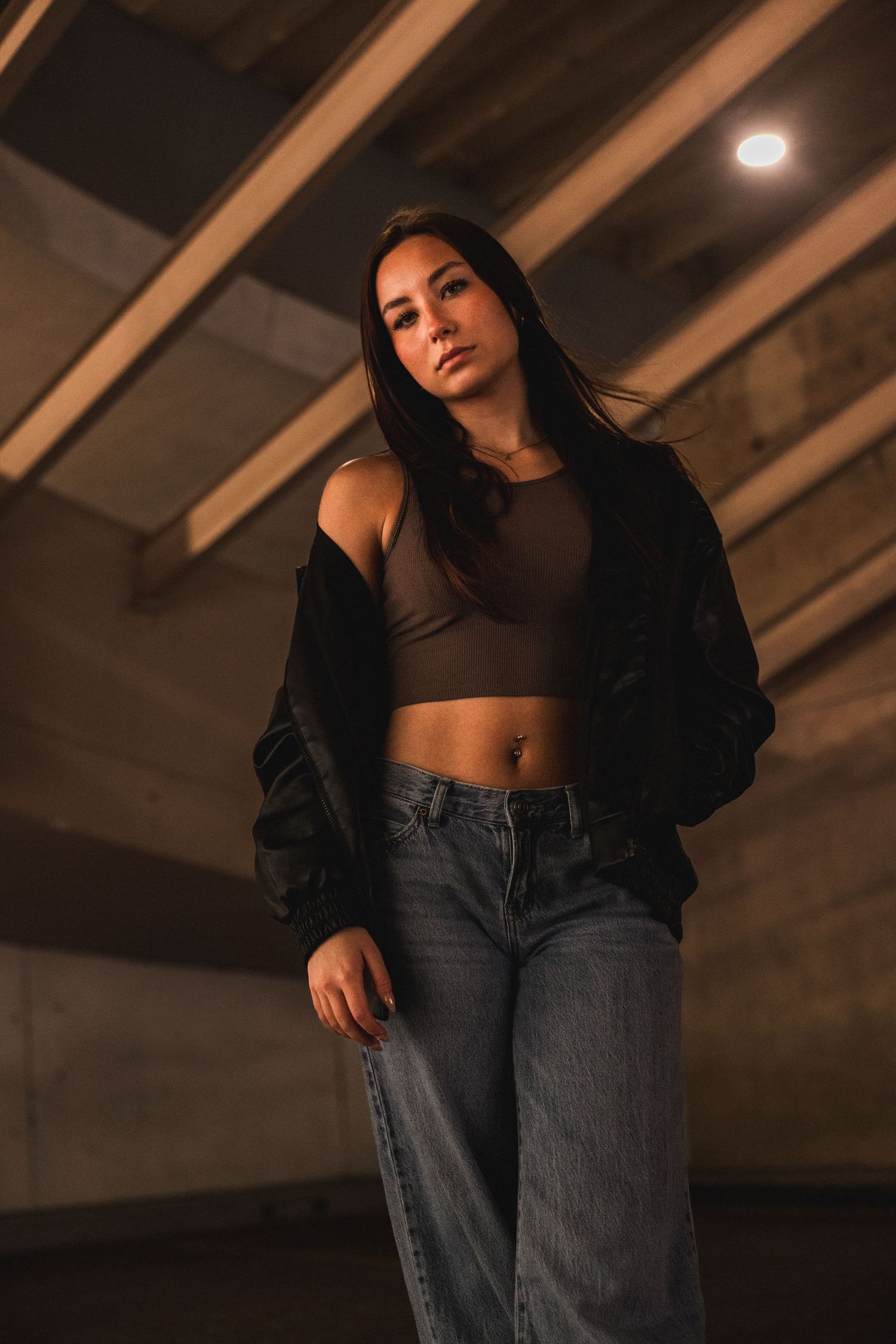 Young woman in crop top, black jacket, and baggy jeans posing confidently in a dimly lit industrial space.