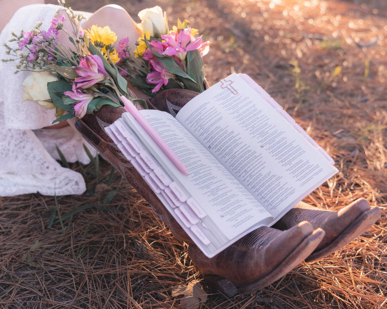 An open book with pink flowers lies on a bed of pine needles in soft natural lighting.