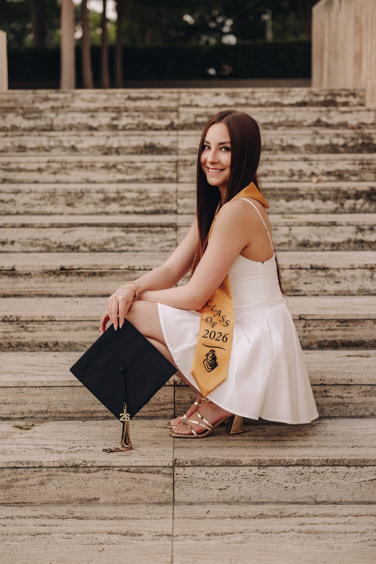 Young woman in white dress holding graduation cap, wearing Class of 2026 sash, sitting on stone steps outdoors.