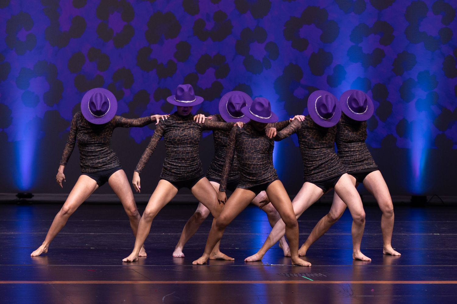 Dancers in black dresses and purple hats perform a synchronized routine on a blue-lit stage.