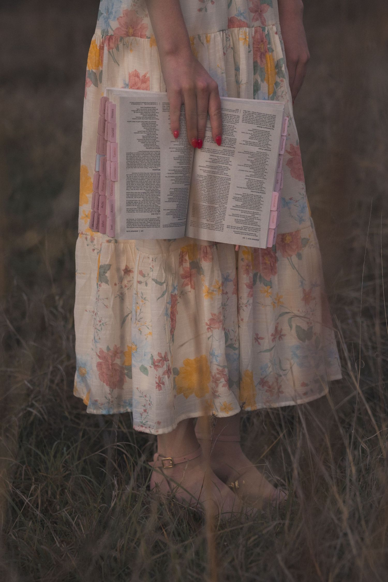 A person in a floral dress holds an open book while standing barefoot in tall grass at dusk.