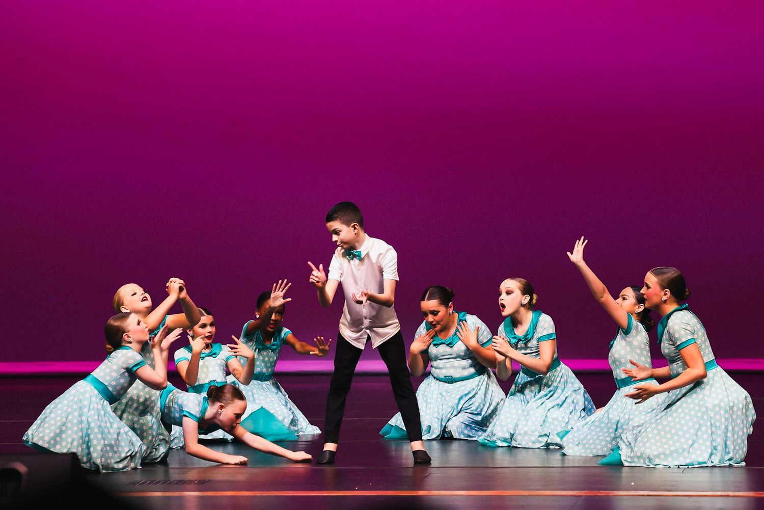 Dancers in blue and white costumes perform a graceful floor choreography on a purple-lit stage.