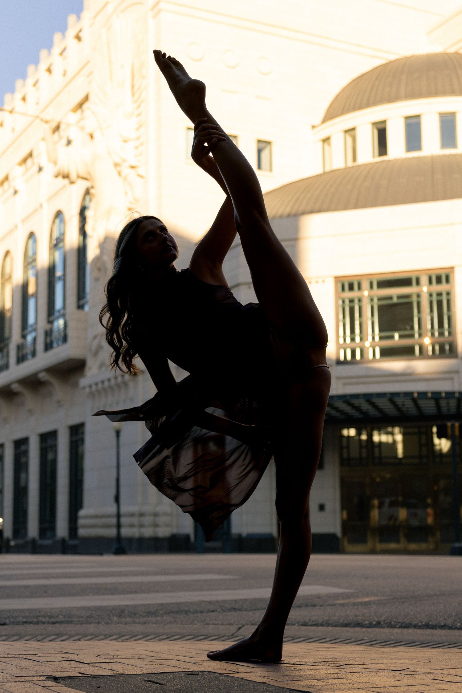 Silhouette of a dancer performing a high leg extension pose outdoors against a sunlit classical building facade.
