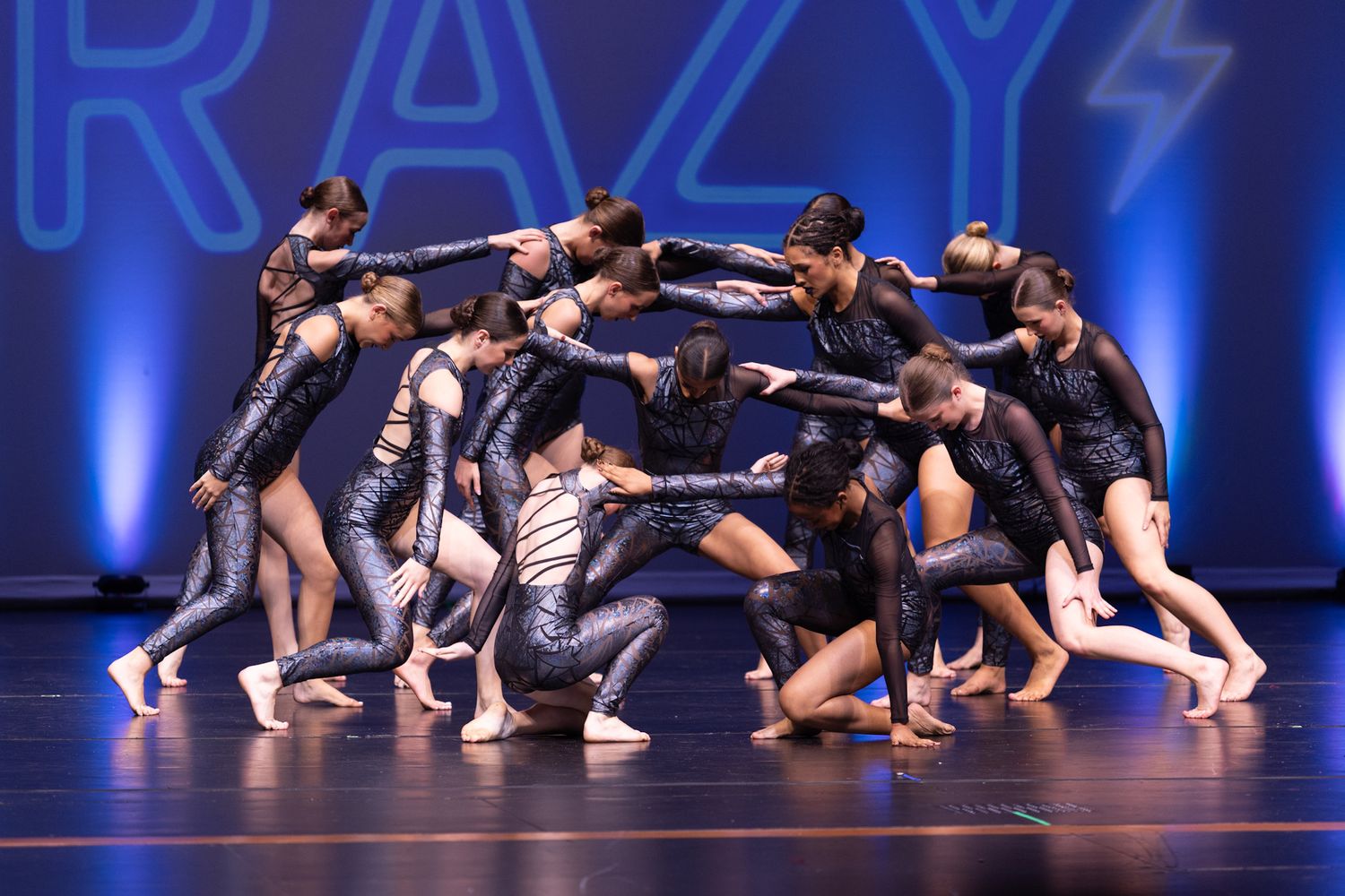 Dancers in sparkly black costumes perform a dramatic group formation on stage with CRAZY backdrop lighting.