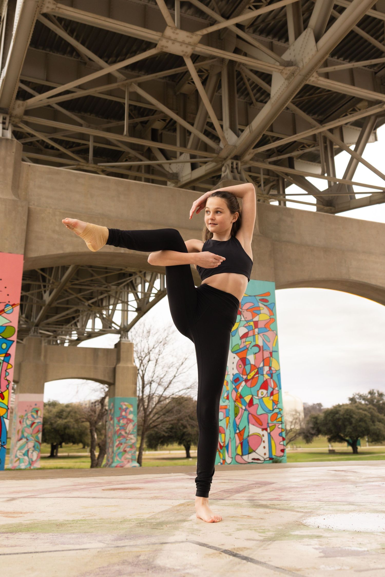 A dancer performs a standing split pose under a concrete bridge decorated with colorful murals.