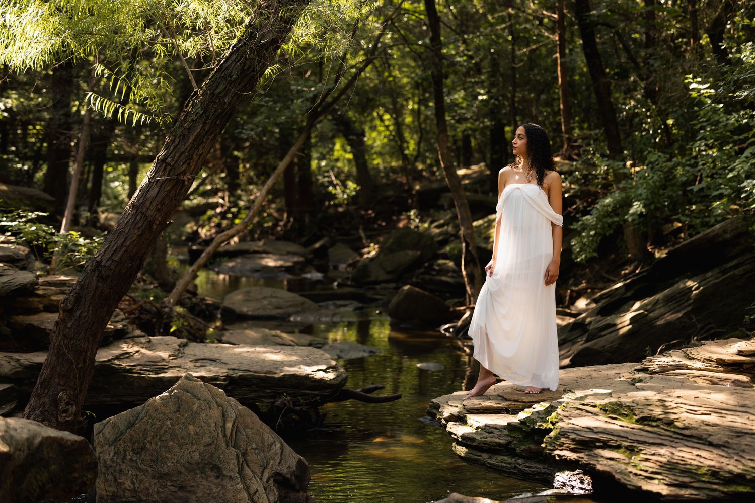 Woman in white dress standing on rocks by a serene forest stream surrounded by lush green trees.