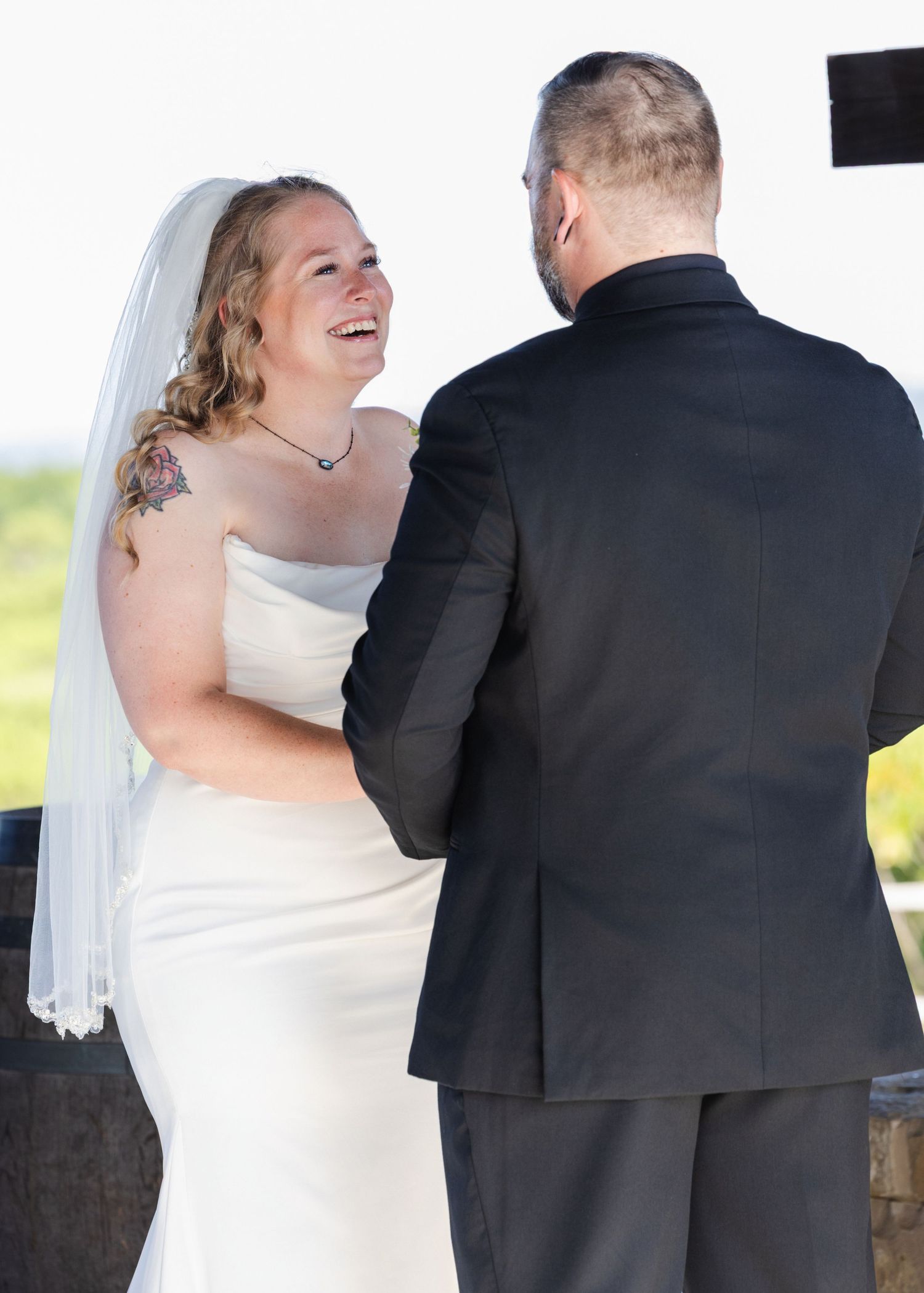 Bride in white gown and veil laughs joyfully with groom in black suit during outdoor wedding ceremony.