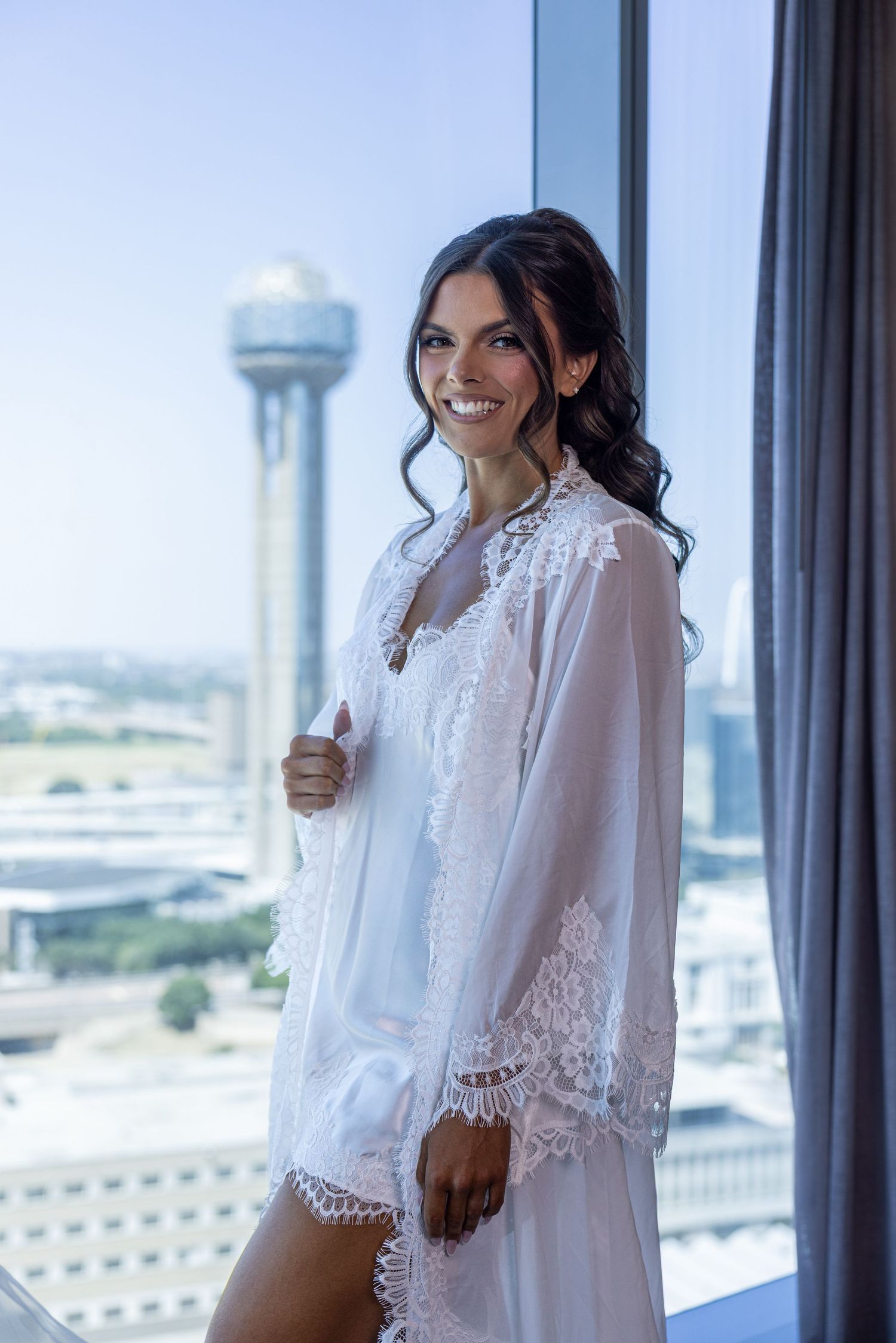 Smiling bride in white lace robe standing by window with city skyline and tower in background.