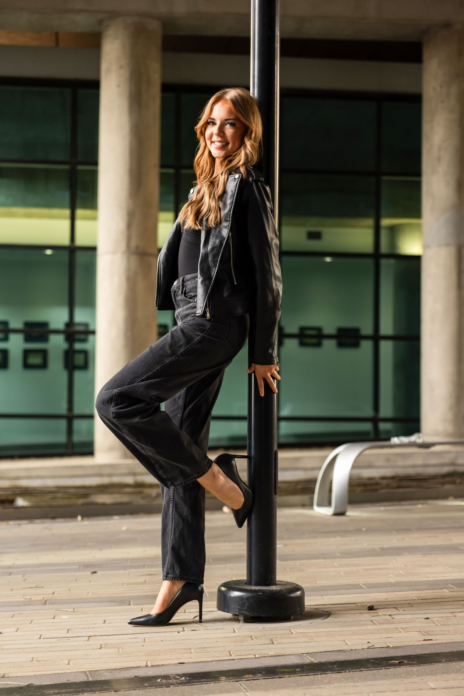 Smiling woman in black outfit leaning against a pole outside a modern building with glass windows.