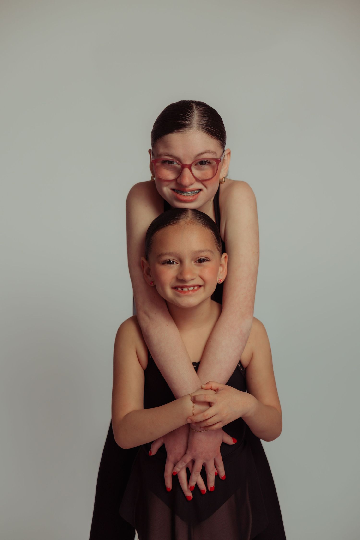 Two smiling girls in dark dresses pose together, older girl with pink glasses hugging younger girl from behind on gray background.