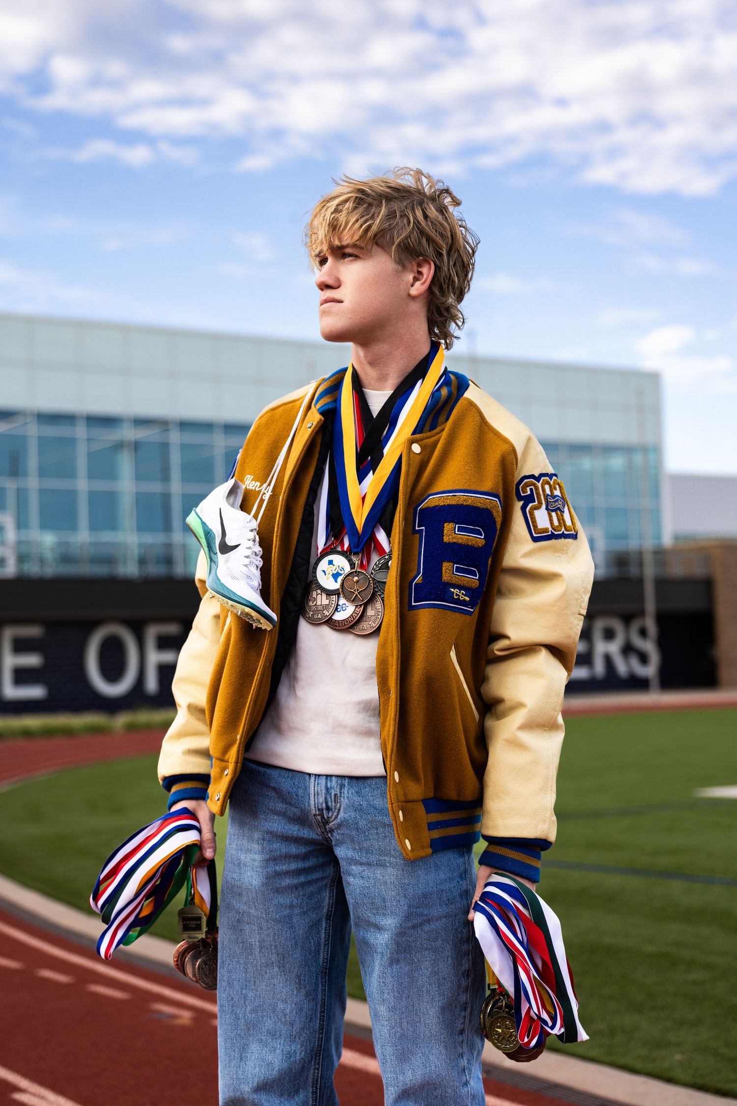 Teen boy in gold letterman jacket wearing multiple medals stands on a track field holding ribbons for a senior portrait.
