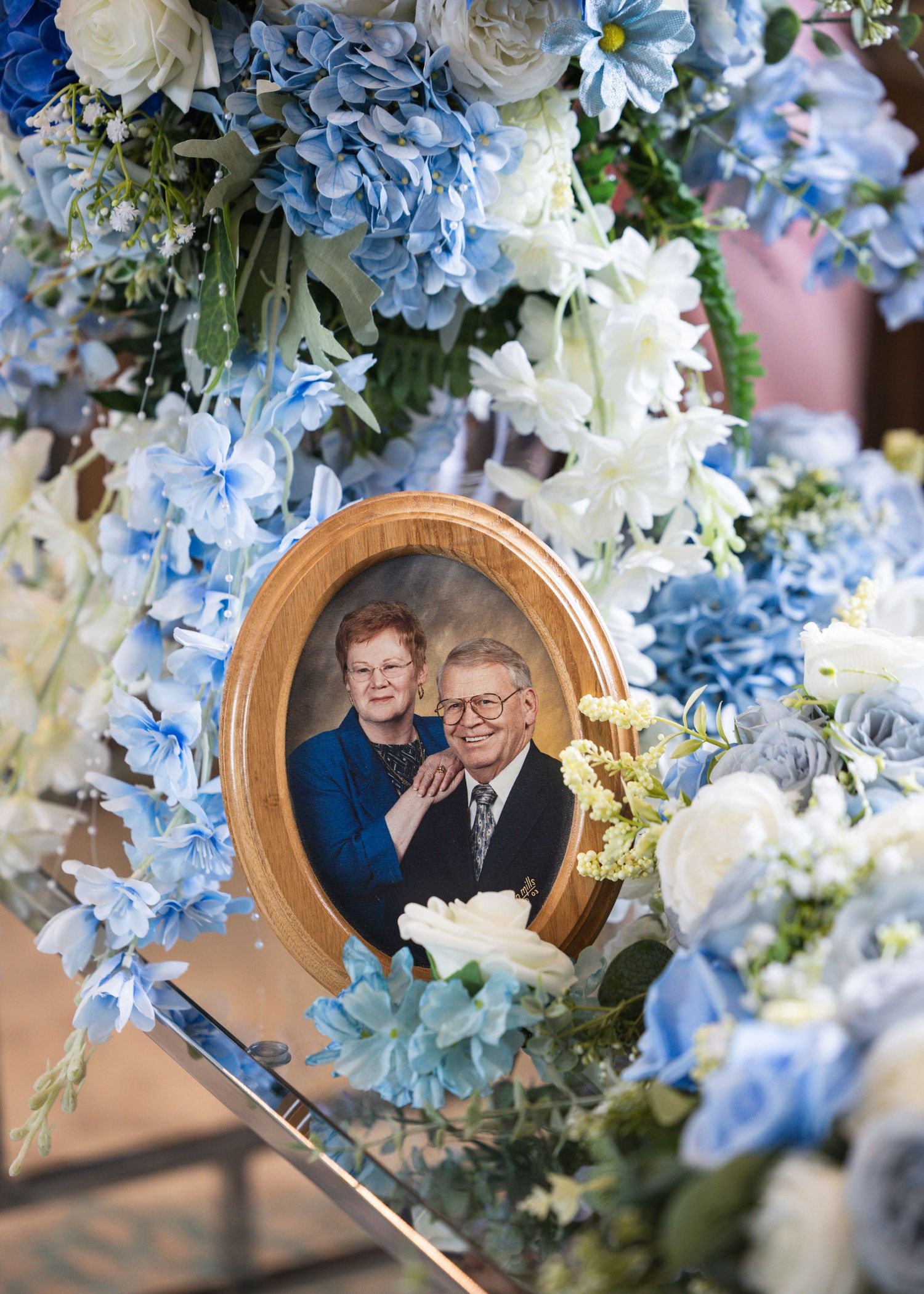 Oval gold-framed portrait of an elderly couple surrounded by blue hydrangeas and white flowers in a floral arrangement.