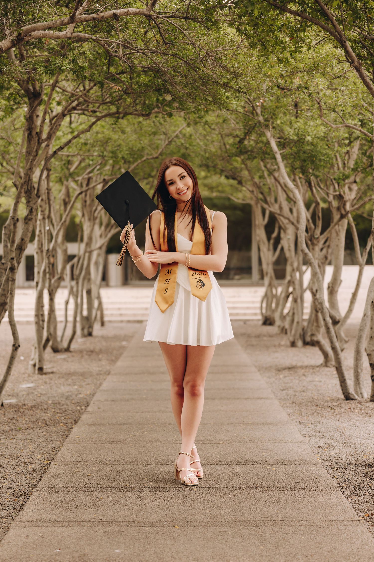 Young woman in white dress holding graduation cap on tree-lined pathway, wearing gold sash for graduation photos.