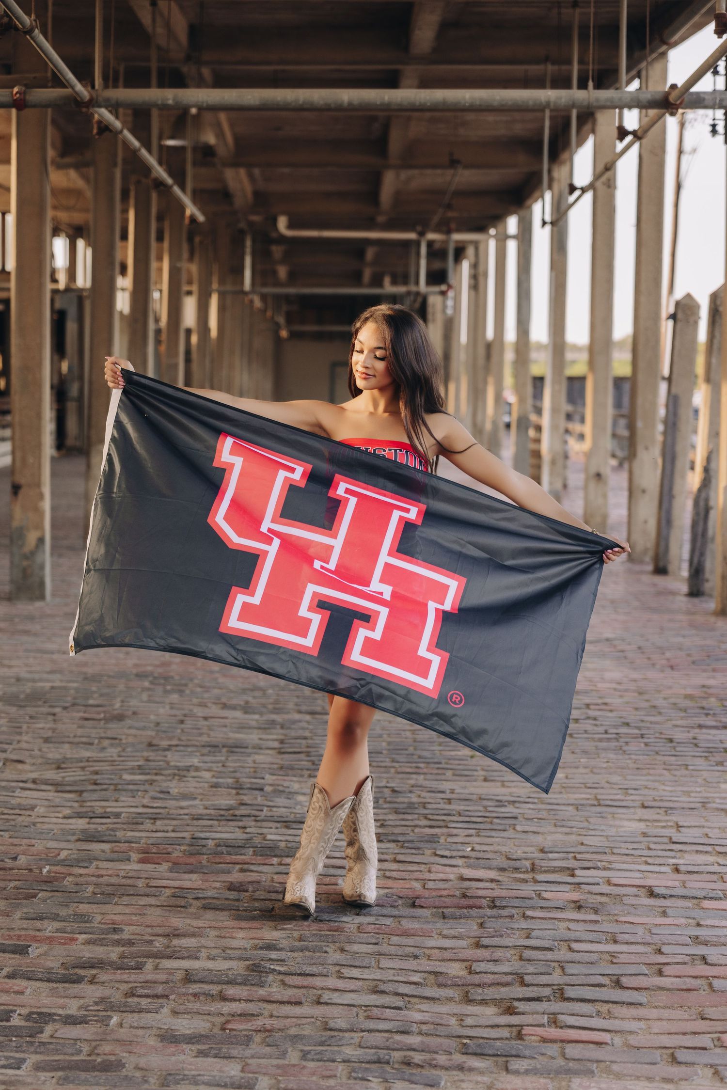 Young woman holding large black University of Houston UH flag wearing cowboy boots on brick walkway.