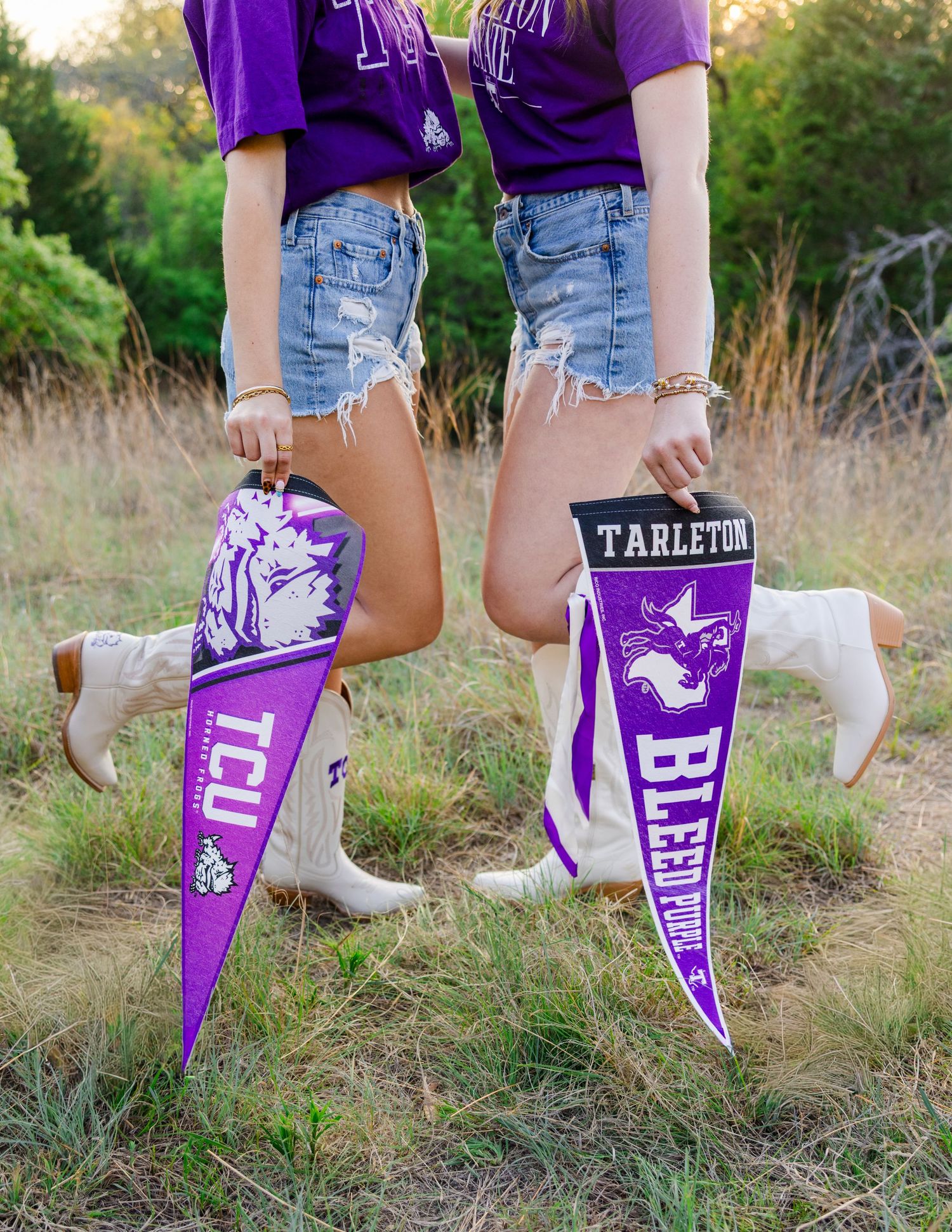 Two girls holding TCU and Tarleton State University pennants in purple, wearing cowboy boots and denim shorts outdoors.