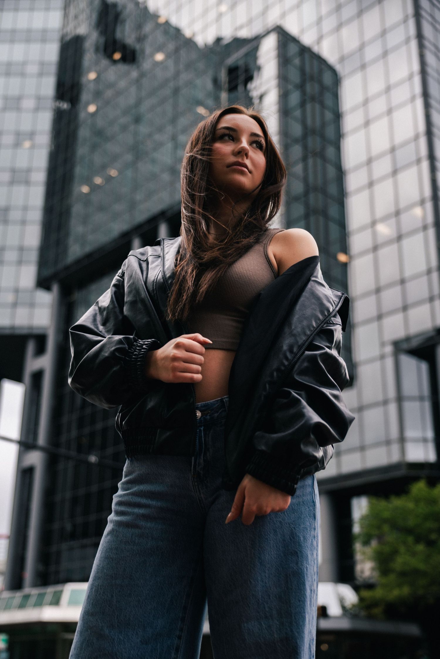 Young woman in leather jacket and jeans poses confidently in front of modern glass skyscrapers in urban setting.