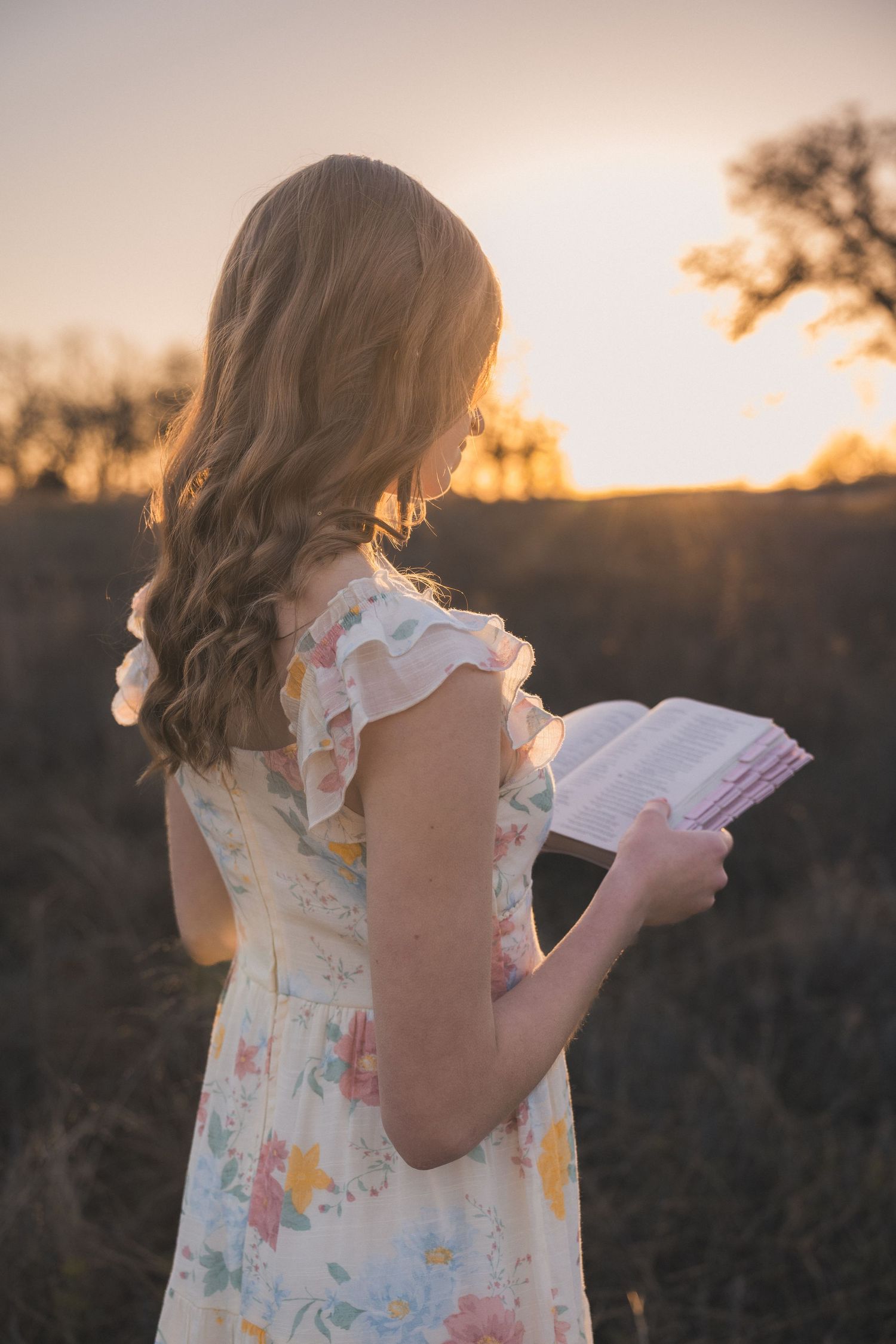 A person in a floral dress reads a book outdoors during golden hour sunset with long wavy hair flowing in the breeze.