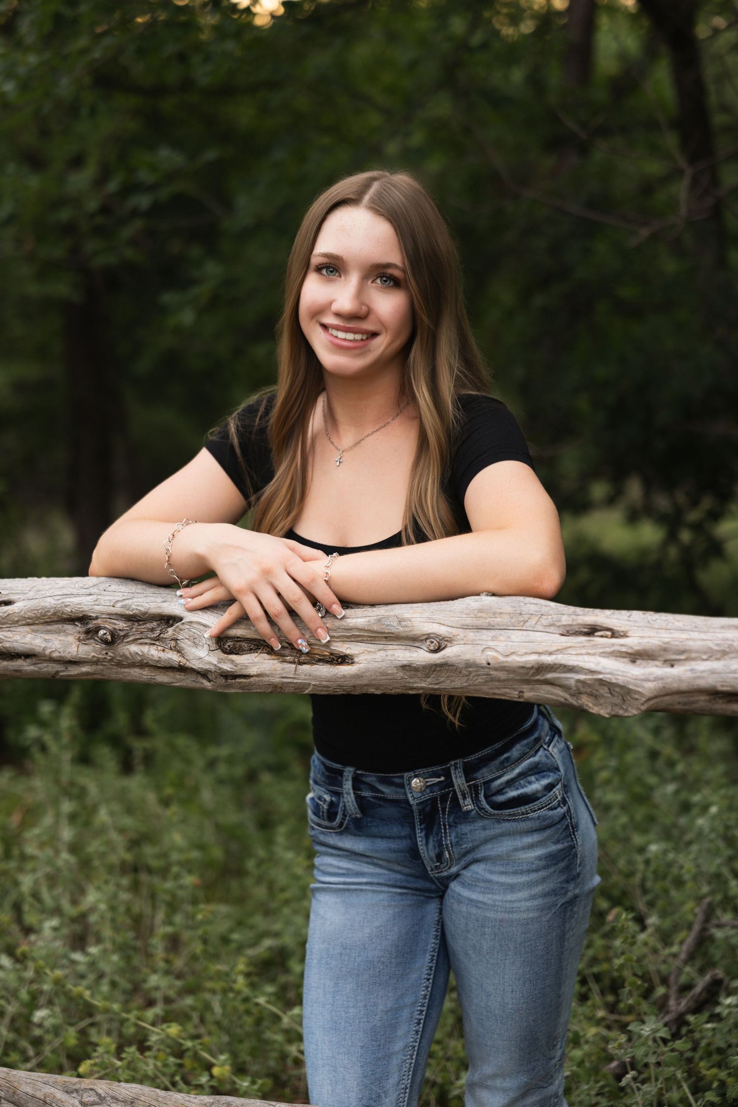 Young woman in black top and jeans smiling while leaning on a rustic wooden log in a lush green forest setting.