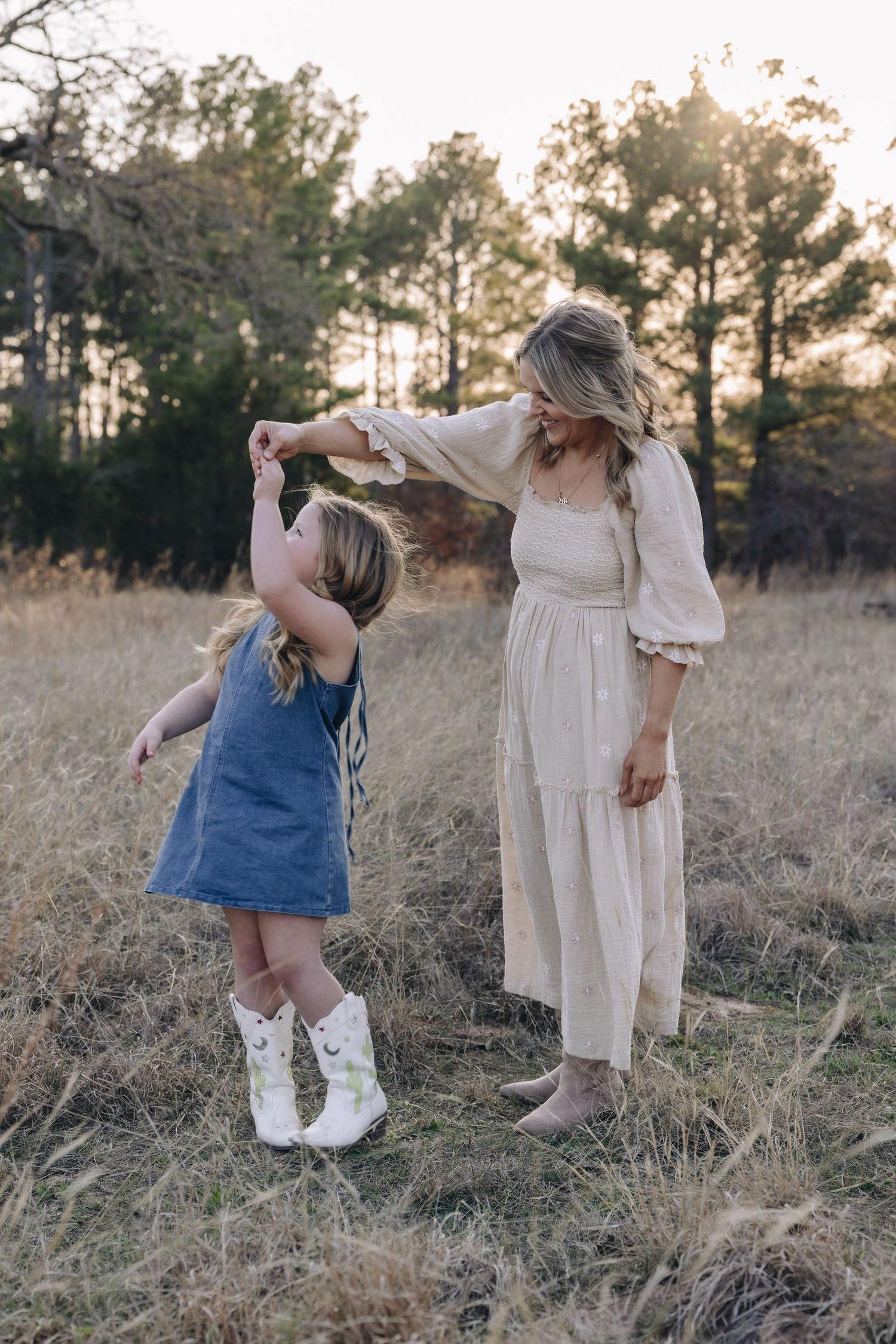 Mother and child dancing together in a grassy field at sunset wearing white and denim outfits.