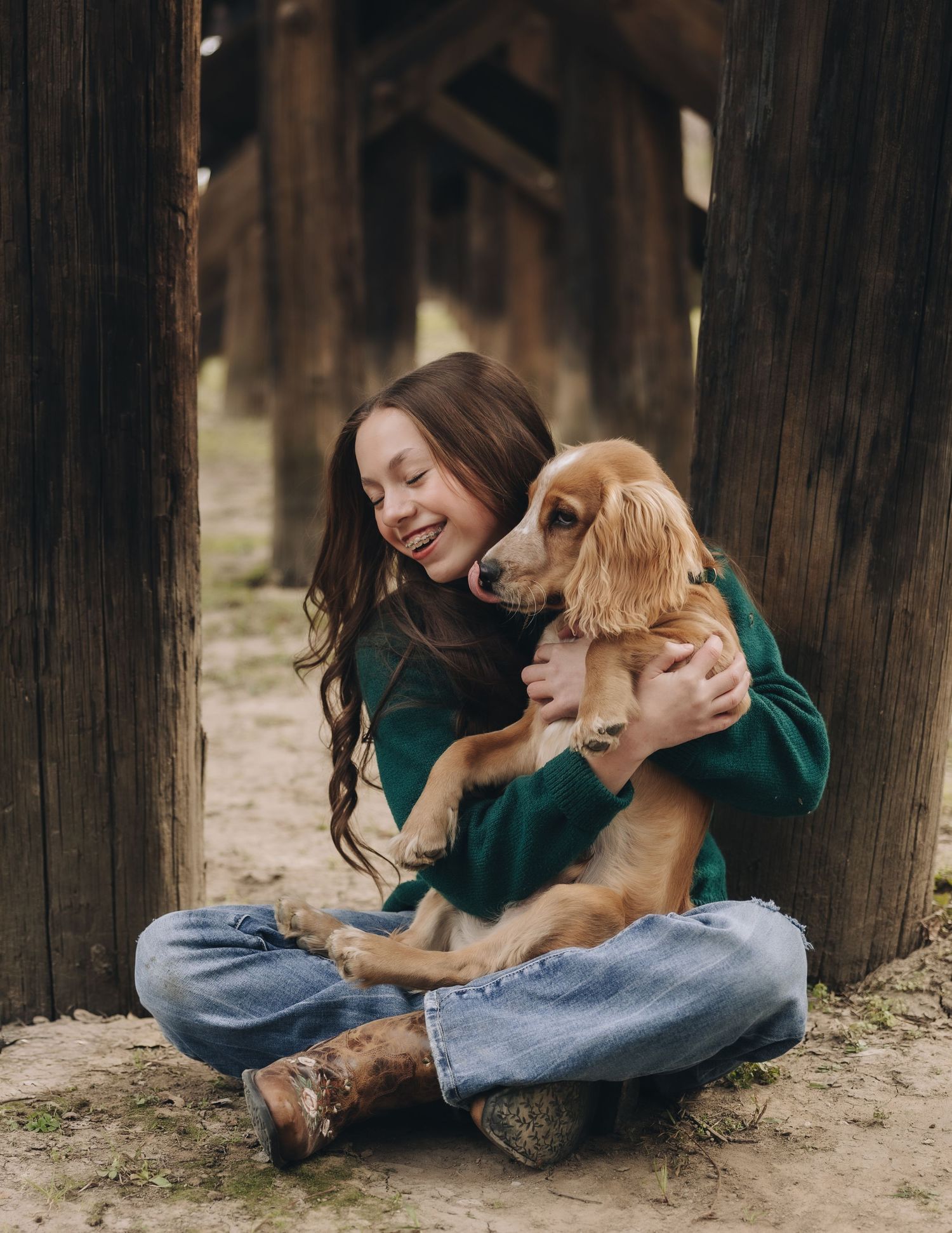 Person in green sweater and jeans sits on ground with golden retriever puppy in forest setting.