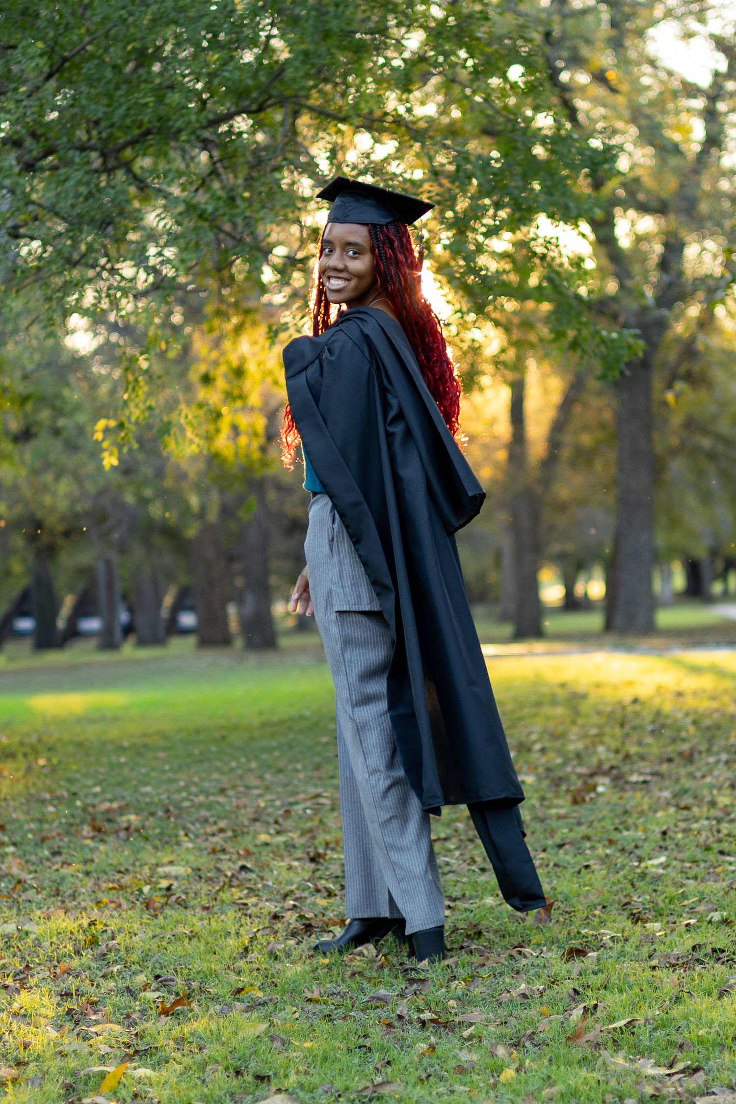 A graduate in cap and gown twirls playfully in a sunlit park with autumn leaves scattered on the ground.